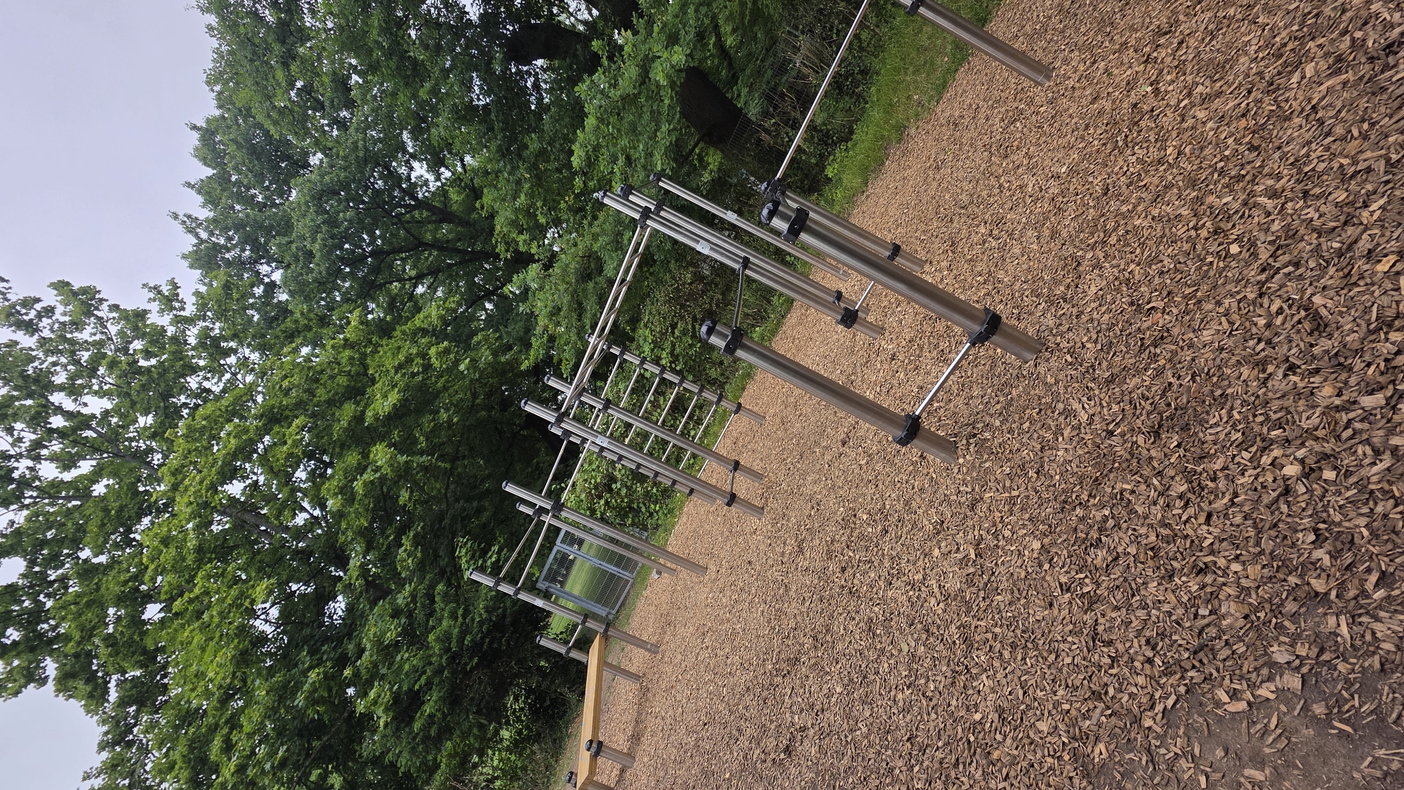 An outdoor fitness area with monkey bars and parallel bars is situated on a ground covered with wood chips. A wooden bench is visible to the left of the fitness equipment. The area is surrounded by trees and foliage, and a metal fence can be seen in the background. The sky is overcast.