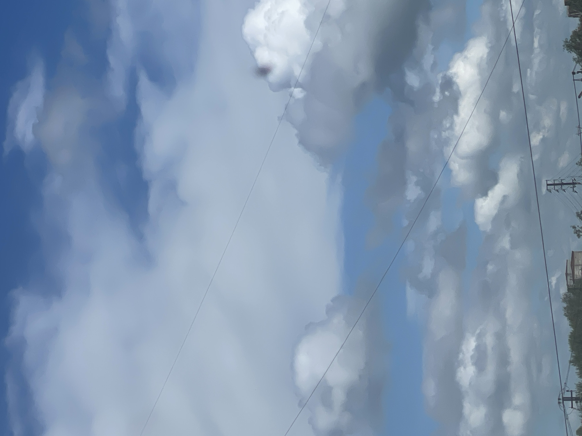 The image shows a view of a cloudy blue sky with power lines stretching across it. In the lower part of the image, there are some trees and structures, possibly buildings and utility poles, visible against the horizon. The clouds are a mix of white and gray, suggesting a partly cloudy or overcast day. The scene appears to be an outdoor environment in a city or suburban area.