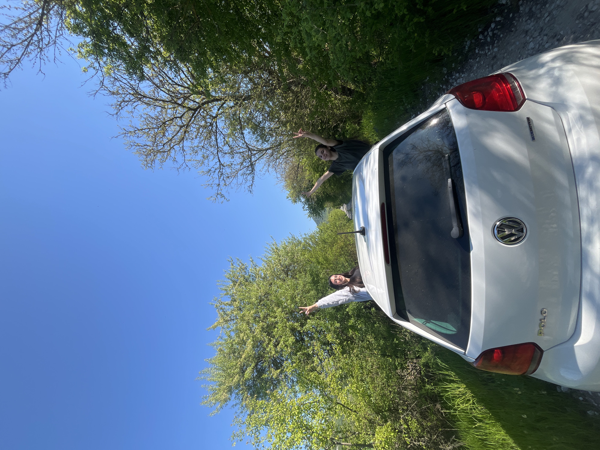 Two people are sticking their arms out of a white Volkswagen Polo car. The car is positioned in front of lush green trees with a clear blue sky above. The photo appears to be taken in a rural area near Rish, Bulgaria. The people are facing towards the car with their arms outstretched towards the trees. One person is wearing a grey sweatshirt and the other is wearing a dark colored shirt. The car is parked on a dirt road near grass.