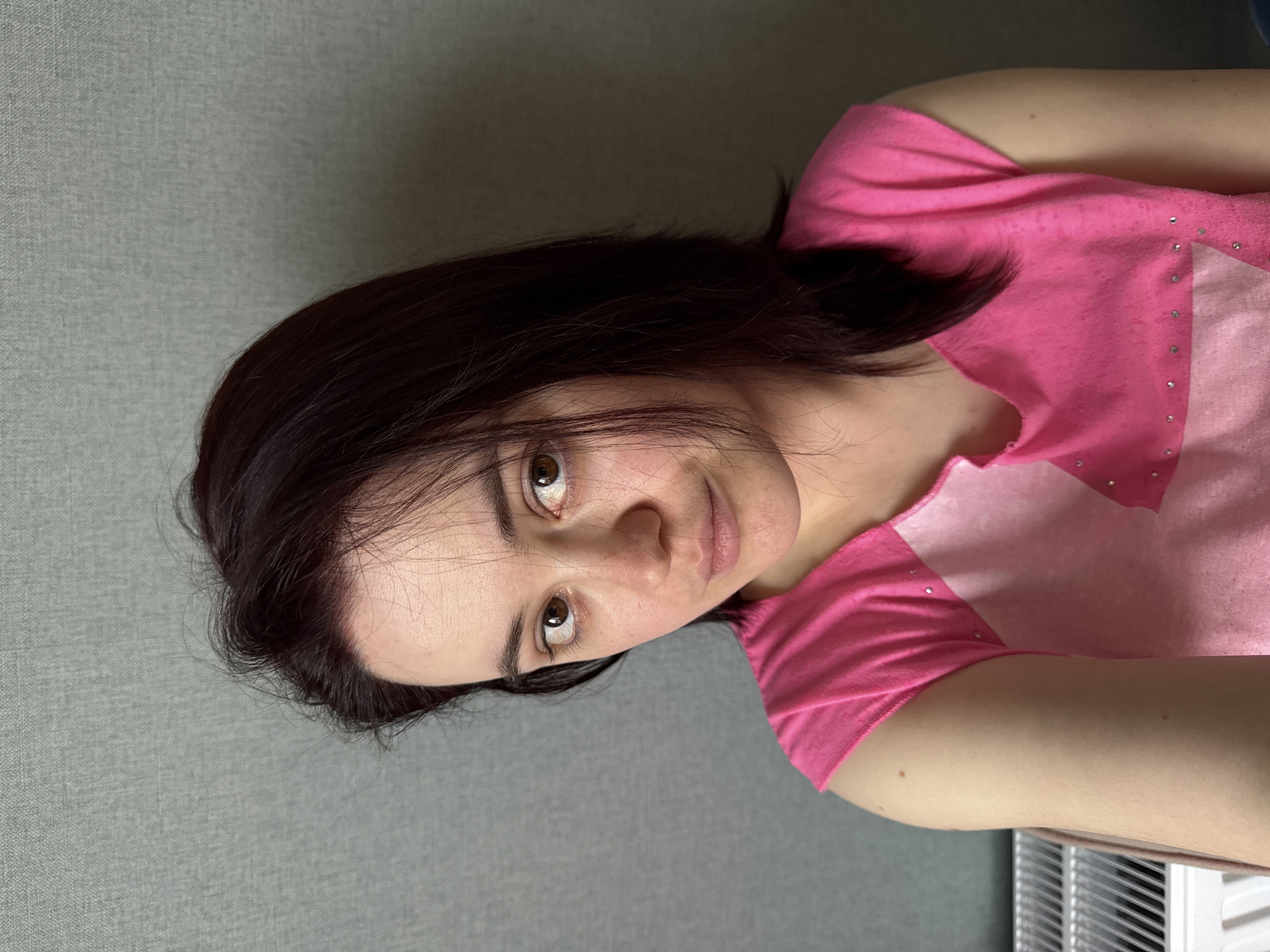A woman with dark hair and brown eyes is shown in a close-up photograph. She is wearing a pink t-shirt with a lighter pink panel on the front, embellished with small rhinestones. The background is a solid gray wall. The setting is indoors. There is no evidence to suggest that this image was taken in the city of Izhevsk, Russia.