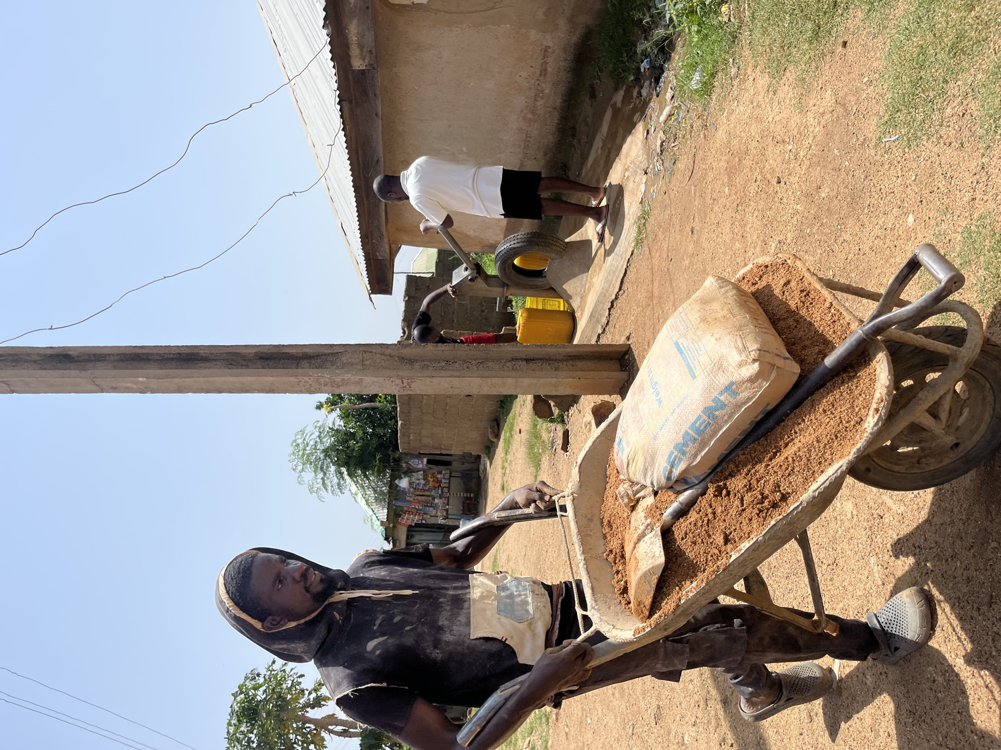 This image depicts an outdoor scene in Jos, Nigeria, with two men engaged in what appears to be construction or maintenance work. In the foreground, a man wearing a dark hooded shirt and dark pants is pushing a wheelbarrow filled with a bag of cement and loose brown material, likely sand. He is wearing grey slip-on shoes. A shovel is visible in the wheelbarrow.
In the background, another man in a white t-shirt and black shorts stands near a building with a corrugated metal roof. He is also near a yellow container and a tire. The setting appears to be a dusty, outdoor area with a building in the background. There are some green grassy patches and some debris scattered around. The lighting suggests it is daytime, likely sunny, given the shadows. The man in the foreground has a focused expression.
Visible text on the bag in the wheelbarrow includes "CEMENT" and possibly "WEIGHT". The overall impression is one of manual labor in a practical, possibly rural or semi-urban environment.