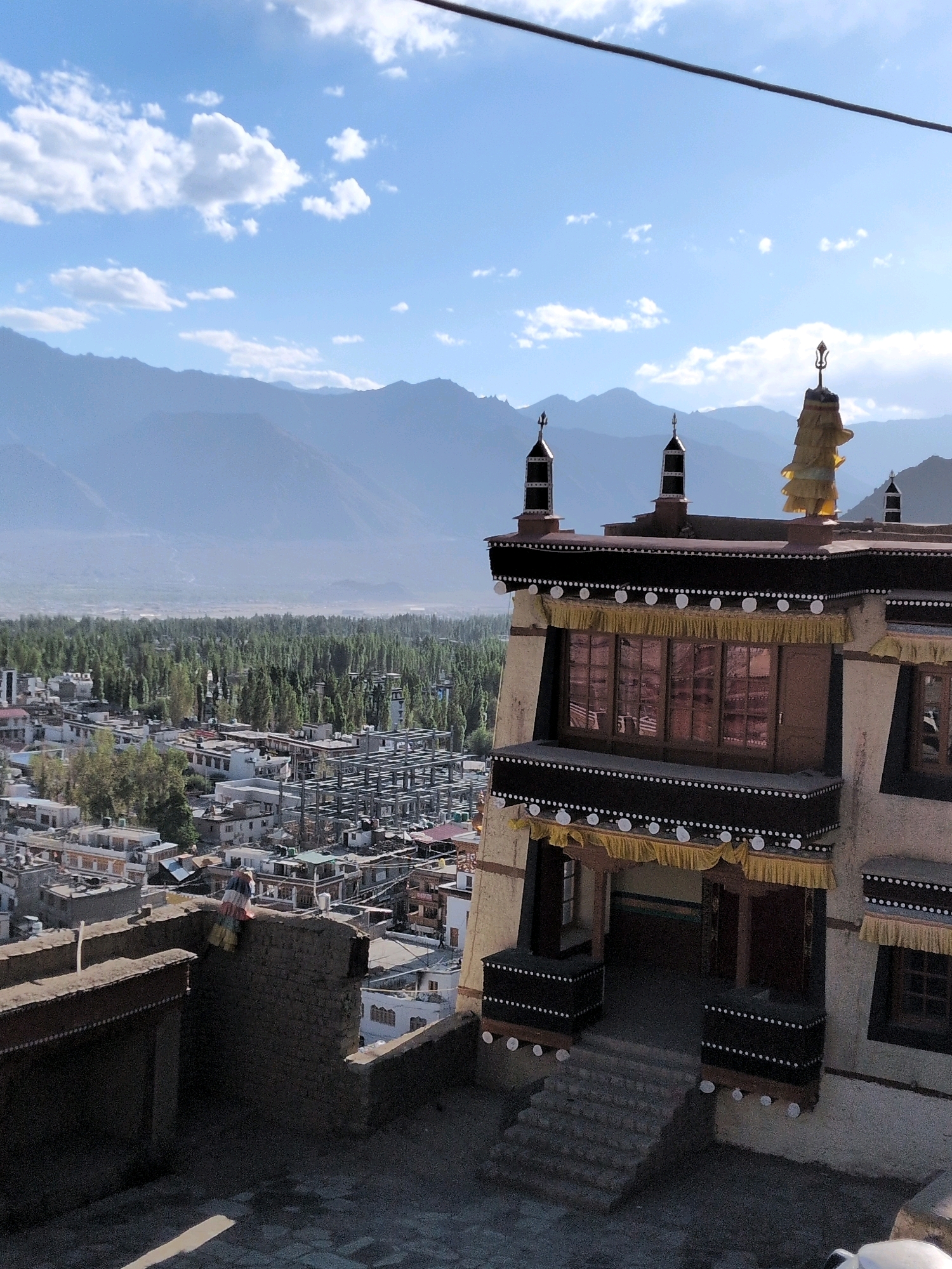 The image shows a view of the city of Leh, India, with a monastery in the foreground. The monastery, likely the Sankar Gompa, is a traditional Tibetan Buddhist structure with whitewashed walls, dark wooden accents, and ornate decorations. It has several levels, with intricately carved windows and doorways. Prayer flags are visible, fluttering in the breeze.
Below the monastery, the city of Leh unfolds, a dense collection of buildings with flat roofs. Many of the structures are small and clustered together, characteristic of an old settlement. A significant number of trees, possibly poplar or willow, are interspersed throughout the city, creating a striking contrast with the arid mountain landscape. In the background, a range of majestic, barren mountains stretches across the horizon. The sky is a clear blue, dotted with a few wispy clouds. The overall scene suggests a bright, sunny day.
In the lower left quadrant, a person is seen seated on a low wall, appearing to rest or observe the surroundings. The person is wearing colorful clothing. The architecture and the landscape strongly suggest the location is Leh, Ladakh, a region in the Indian Himalayas known for its Buddhist monasteries and dramatic mountain scenery.