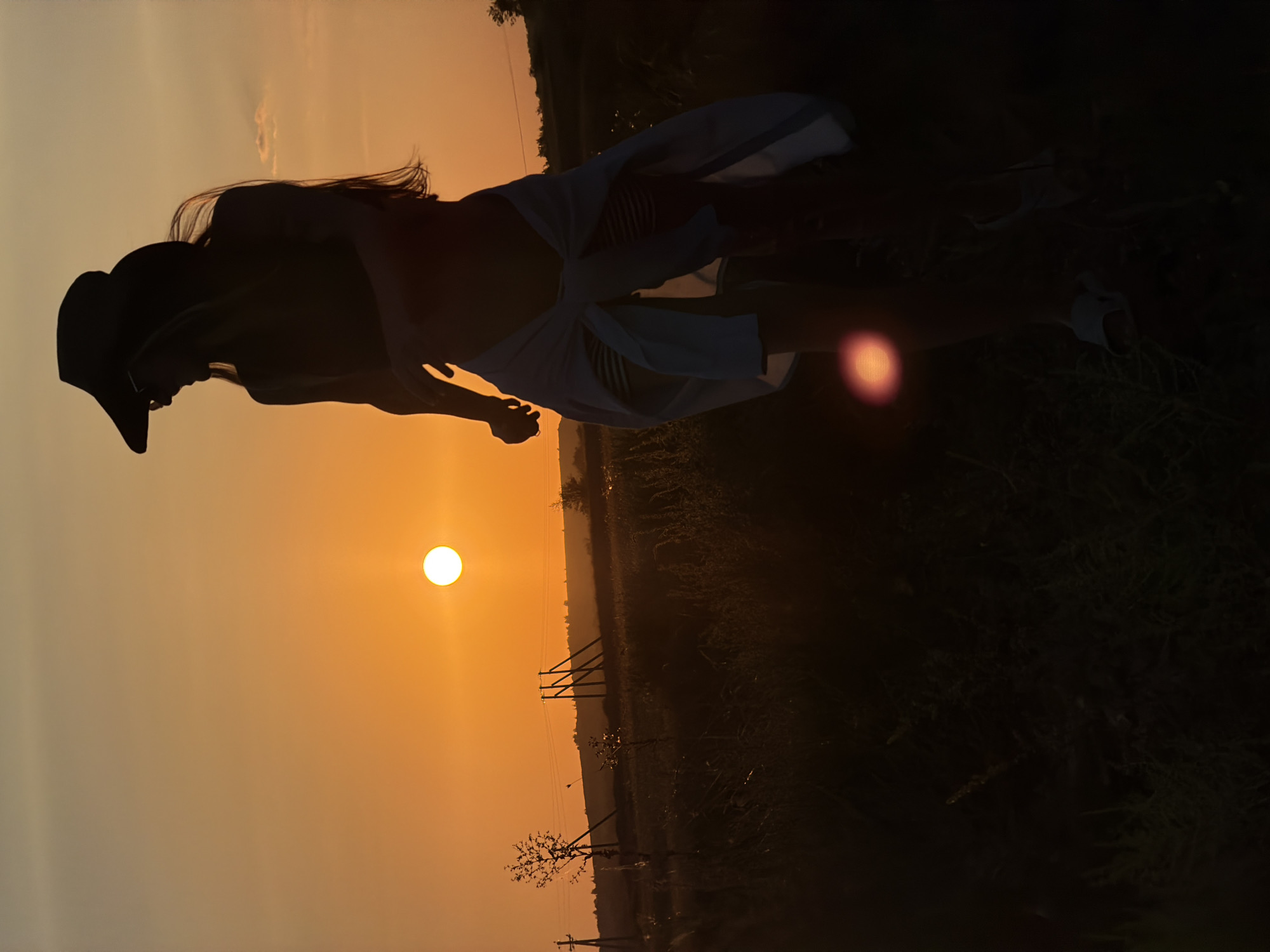 A full-length, silhouetted shot features a person wearing a wide-brimmed hat, sunglasses, and a white shirt tied around their waist as they walk through a field at sunset. The sun is a bright, circular orb in the orange sky. The person's long hair flows behind them. The landscape consists of dry grass and sparse trees, with power lines visible in the distance. The location is stated to be Kamyshin, Russia.