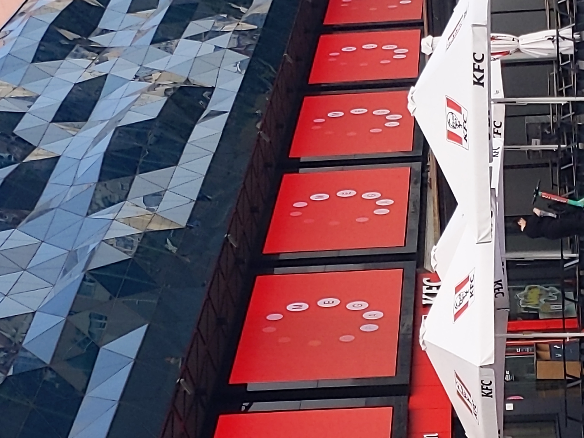 The image depicts the exterior of a KFC restaurant, likely in Kyiv, Ukraine, given its distinct architecture. The building features a modern facade with a geometric pattern of dark, reflective panels on its upper section. Below this, a series of large, bright red digital screens display white circular elements, with some screens showing letters. In the foreground, two white KFC branded umbrellas are set up, providing shade over outdoor seating areas. The KFC logo, featuring Colonel Sanders, is prominently displayed on the umbrellas. A person in dark clothing is visible in the middle ground, standing near a scooter, suggesting the presence of pedestrian traffic. The overall scene conveys a commercial urban environment, likely a busy street or public area.
