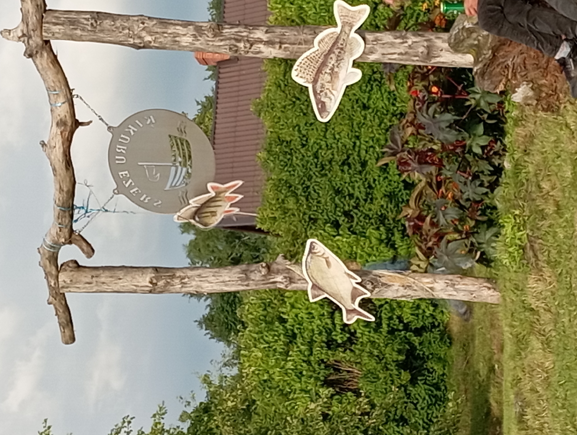 A wooden signpost with a horizontal beam made of branches supports a circular metal sign with a boat and waves. Two paper cutouts of fish are attached to the signpost, one to the left and one to the right. The signpost is located outdoors, with lush green foliage in the background. The ground is covered with grass and some plants. A small section of a brown tiled roof is visible behind the foliage. A person's legs are visible in the lower right corner of the frame, wearing dark pants and shoes, and holding a green can. The sky is overcast.