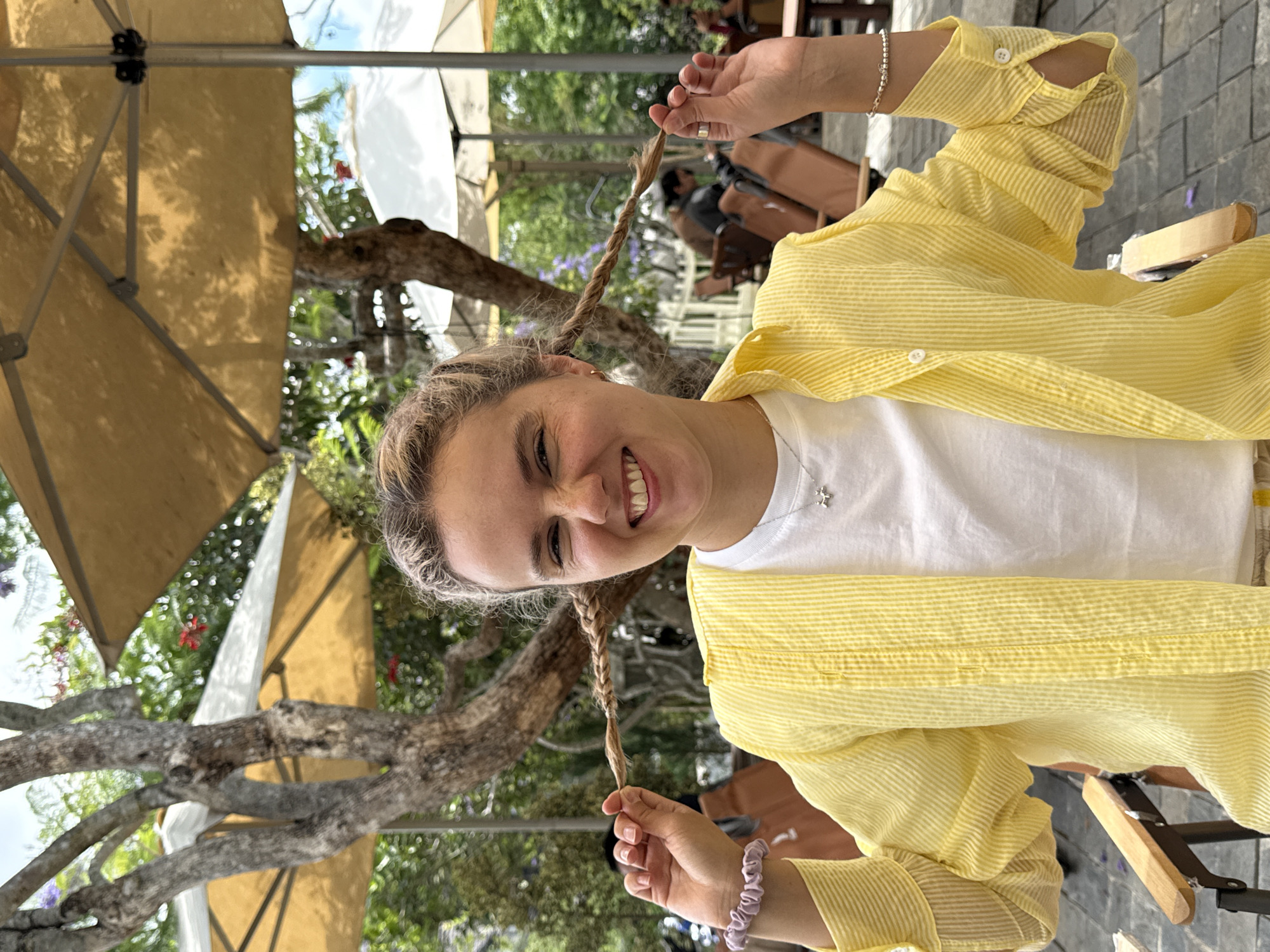 A smiling woman is depicted in a tilted, medium-shot outdoor photograph, likely taken in Da Lat, Vietnam, given the surrounding elements. She is wearing a white t-shirt underneath a striped yellow button-down shirt. Her hair is styled in two braids, which she is holding up with her hands. A silver bracelet is visible on her right wrist, and a lilac-colored scrunchie adorns her left. She also wears a necklace with a cross pendant.
The background includes outdoor seating, beige umbrellas, and trees, indicating she is at a cafe or similar establishment. Other people are present in the background. The location implies a public space in Da Lat, known for its outdoor cafes and pleasant climate.