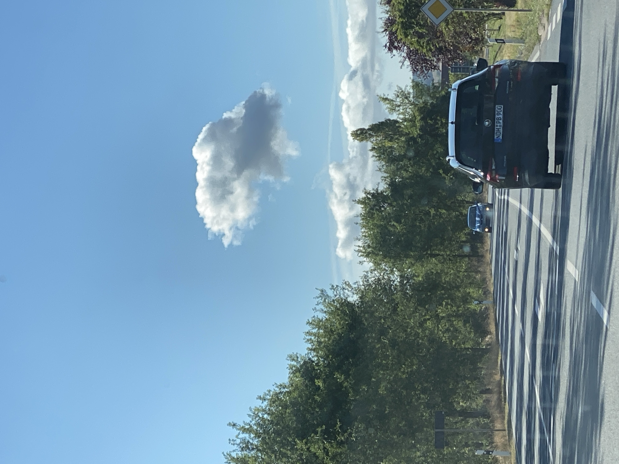 A black SUV with license plate NDH PB 900 is traveling on a two-lane road in Nordhausen, Germany, on a clear, sunny day with a few clouds in the sky. The SUV is in the right lane, followed by another car in the distance. The road is lined with trees on both sides. A yellow diamond-shaped sign is visible on the right side of the road. The image appears to be taken from another vehicle.