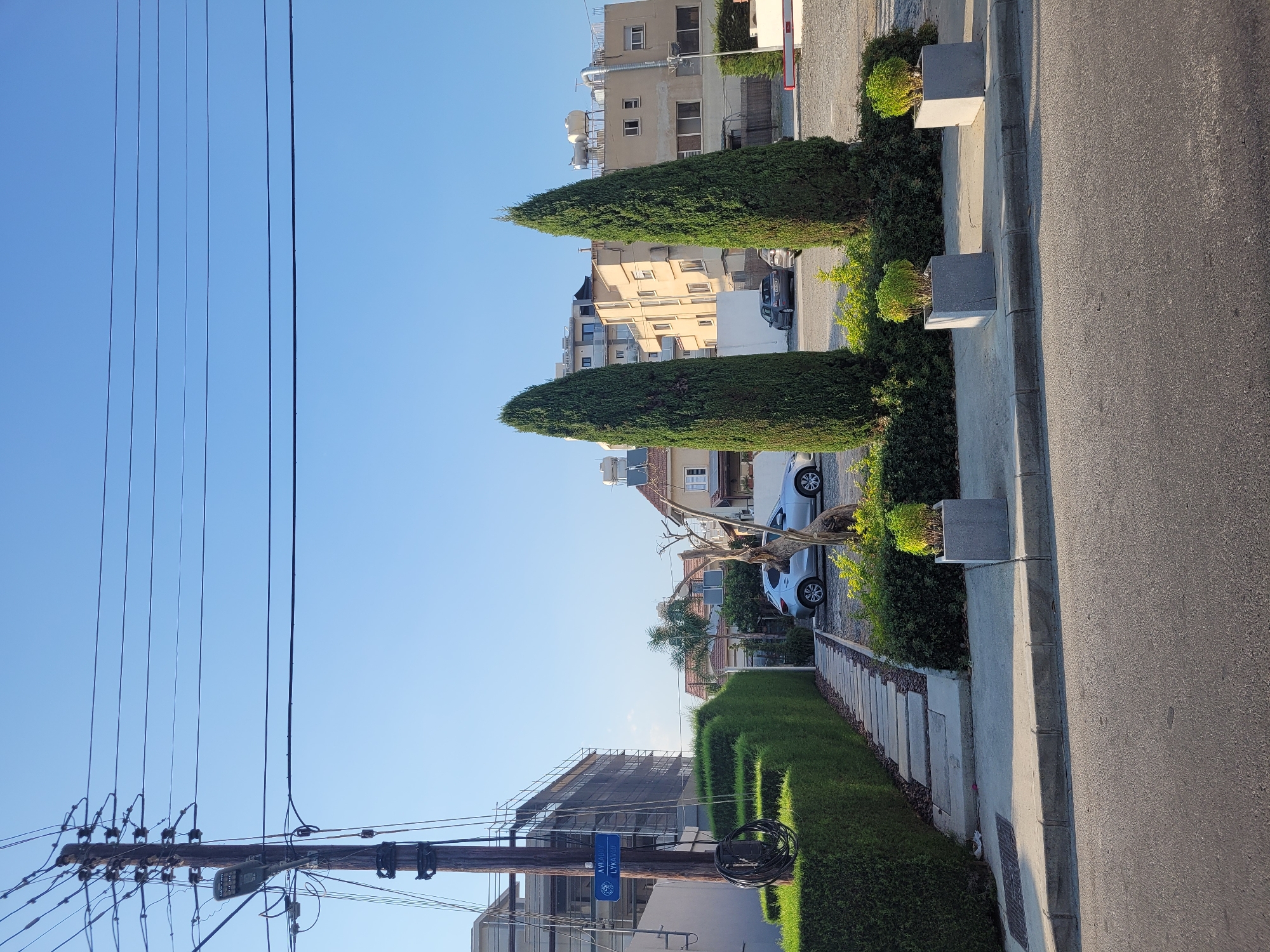 The image depicts a street scene in Limassol, Cyprus, under a clear blue sky. In the foreground, a paved road runs horizontally. To the left, a well-maintained hedge forms a border along a concrete pathway that leads to a property. A wooden utility pole with numerous wires is prominently positioned on the left side of the frame, displaying a blue street sign that reads "AYKABY LYKAVOU." Behind the hedge and pathway, a silver car is parked. Further back, a modern building is under construction, covered in scaffolding. Two tall, conical cypress trees stand in the middle ground, flanked by more greenery and parked cars. In the background, residential buildings with multiple stories and balconies are visible. Several rooftop water tanks and air conditioning units are also present on the buildings. The overall scene suggests a residential or mixed-use area in a Mediterranean urban environment.