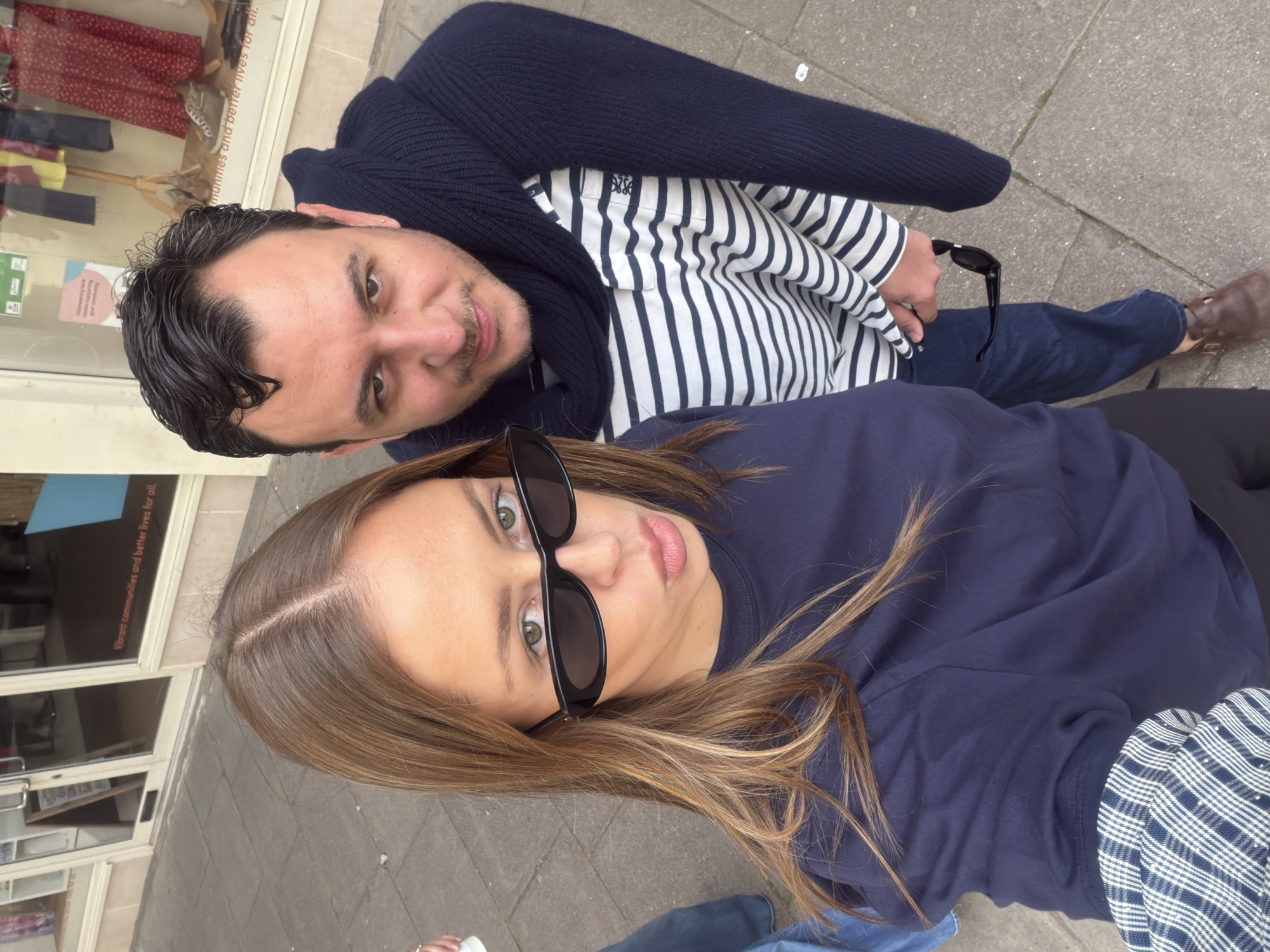 Here is a detailed description of the image:
**People:** The image features two individuals, a man and a woman, who are taking a selfie.
The man has dark hair combed back, and a trimmed mustache. He is wearing a white shirt with navy blue stripes, covered by a navy blue sweater or jacket.
The woman has medium-length brown hair. She is wearing large black sunglasses and a dark blue top.
**Setting:** The setting appears to be an outdoor urban environment, likely a street or sidewalk. A store front is visible in the background, with visible text on the window including "communities" and "lives for all.". The pavement is visible beneath the subjects.
**Objects:** The prominent objects include the sunglasses worn by the woman, clothing worn by both individuals, and the possible presence of a smartphone used to take the selfie.
**Actions and Interactions:** The primary action is the taking of a selfie. The man and woman are posing closely together, indicating a personal relationship.
**Overall Impression:** The photo is a candid shot of two people in an urban setting, possibly a street in the city of London, United Kingdom given the text on the window.