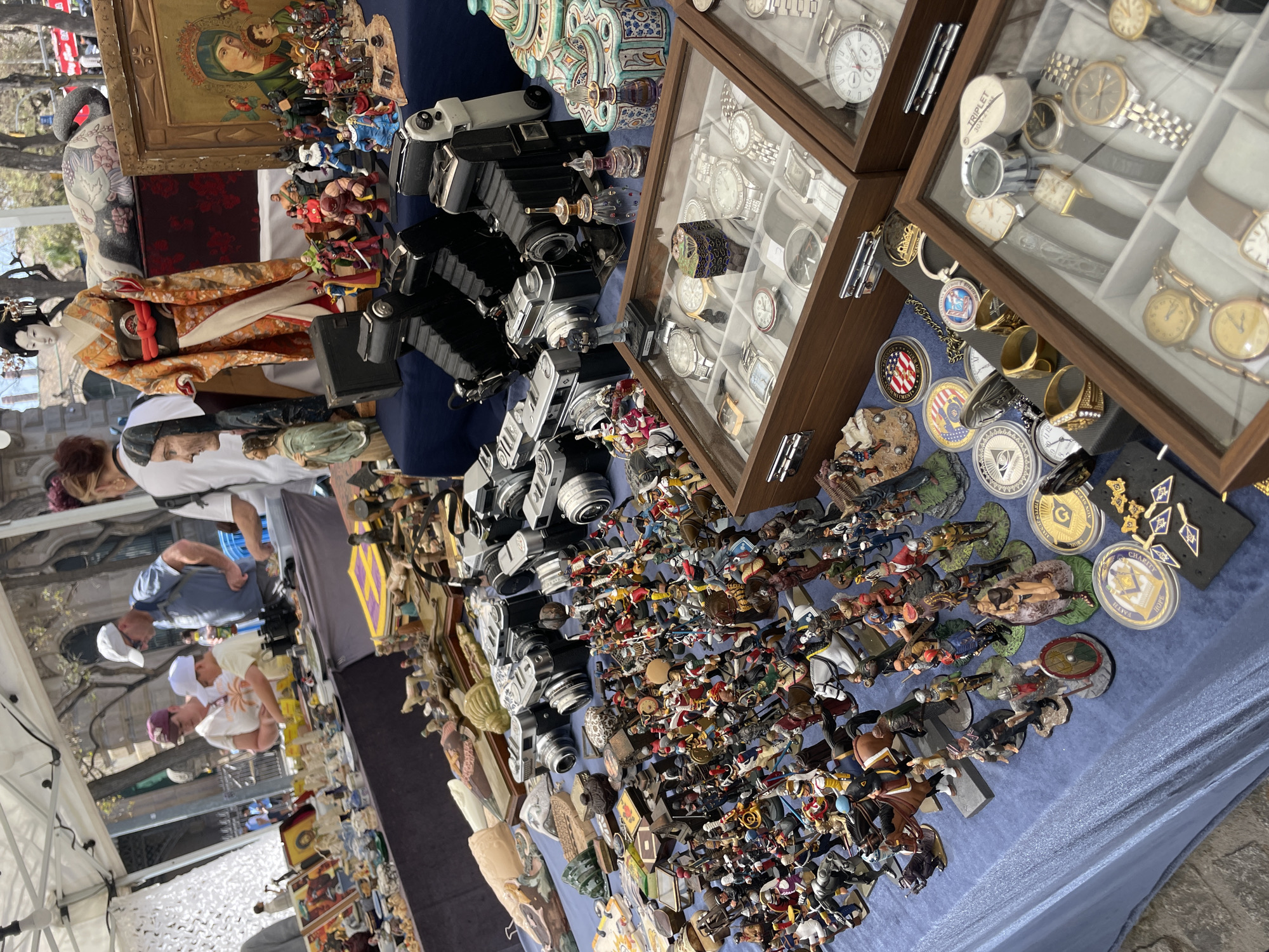 This image shows a vendor's table at a market in Barcelona, Spain. The table is covered with a blue cloth and features a variety of vintage items for sale including vintage cameras, watches displayed in wooden cases, collections of small figurines, coins, and decorative items. Several customers are browsing the items.