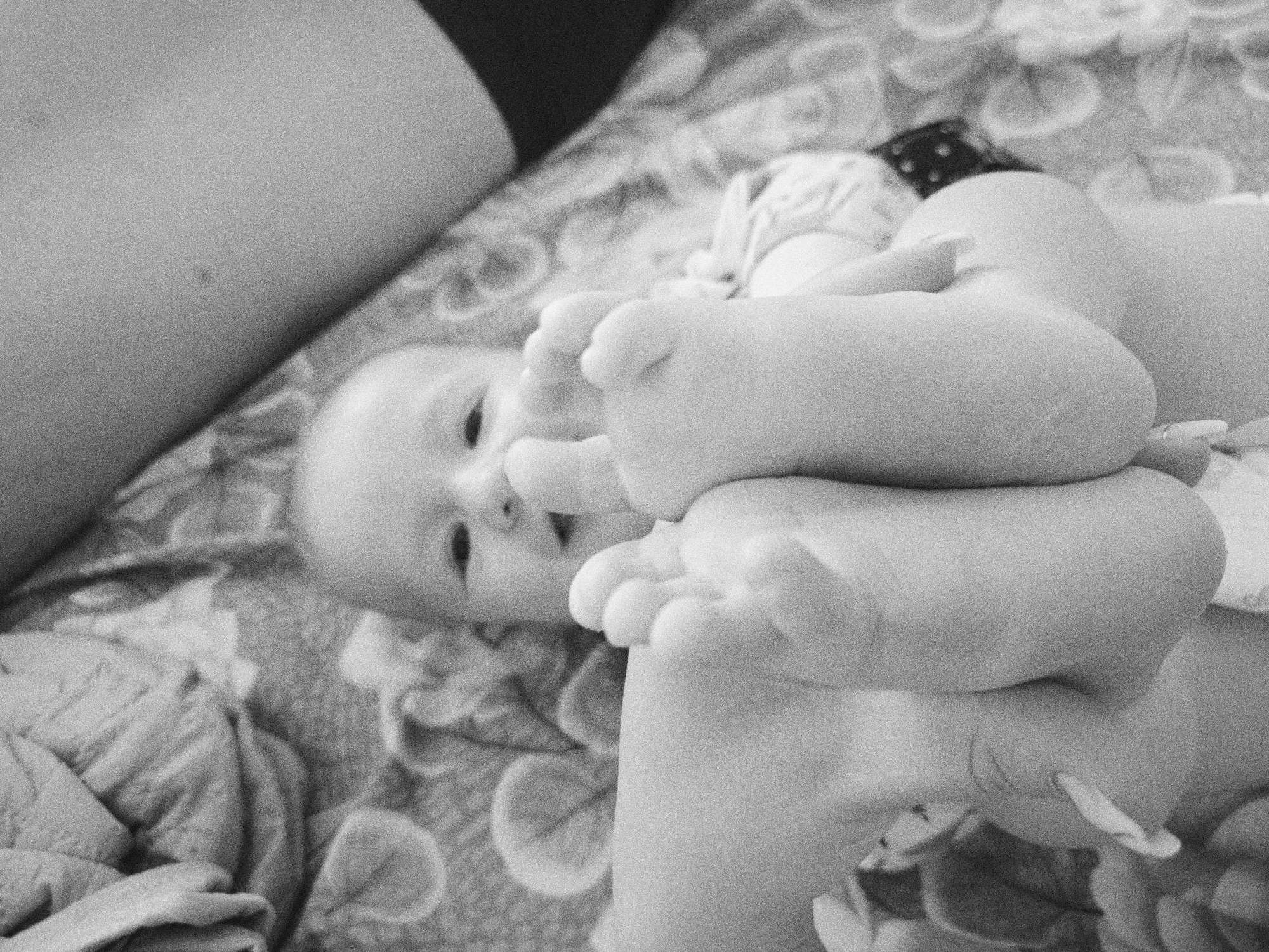 A black and white close-up shot depicts a baby lying on a patterned bedspread, holding its feet. The baby's face is visible, and an adult's arm or leg is also in the frame. There's no information to suggest the image was taken in Tobolsk, Russia.