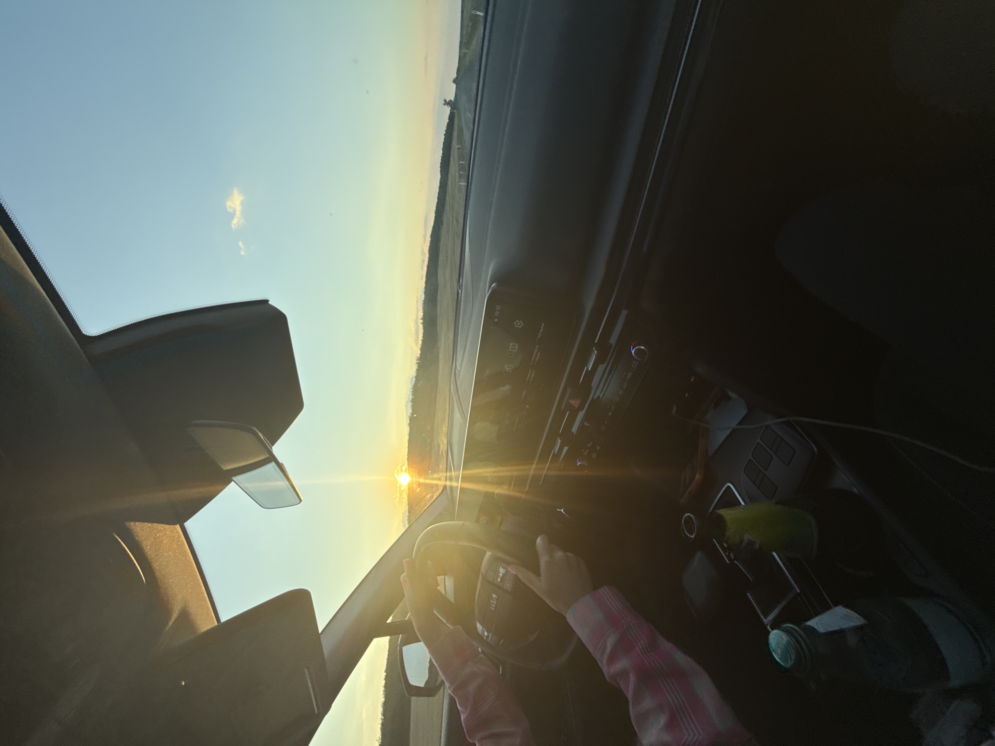 This is a photograph taken from inside a vehicle, looking towards the dashboard and out the windshield at sunset. The sun is low on the horizon, casting a warm glow and lens flare across the scene. A person's hands are visible on the steering wheel, wearing a pink and white checkered long-sleeved shirt. The interior of the car includes a modern dashboard with a central infotainment screen displaying icons and text. Two bottles are visible in the center console, one green and one clear, along with a charging cable. The exterior view shows a rural landscape with rolling hills and a road stretching into the distance. The sky is a clear blue with a few wispy clouds. The photograph's perspective and the presence of a road suggest it was taken while the vehicle was in motion, possibly on a highway or country road outside of Sadaclia, Moldova.