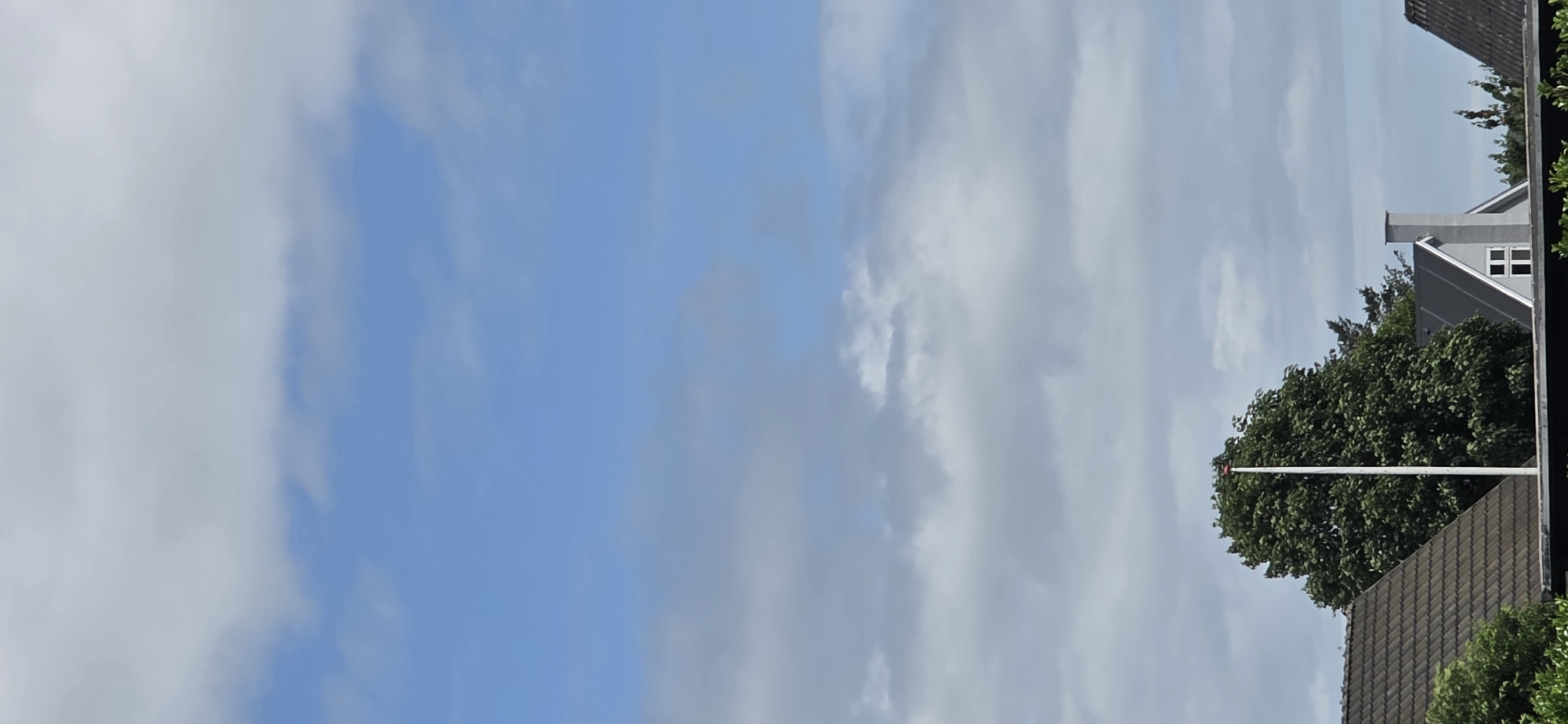 This is a vertical, outdoor, full shot image taken in Hjørring, Denmark. The upper two-thirds of the image are dominated by a blue sky with scattered clouds, some of which appear to be cumulus. The bottom third of the image shows the rooftops of houses and a large, green deciduous tree. A tall, white flagpole with a red cap stands prominently in front of the tree. The roofs are tiled, with one house featuring a gray facade and a chimney with a white window frame. The overall impression is a clear, partly cloudy day with residential buildings visible.