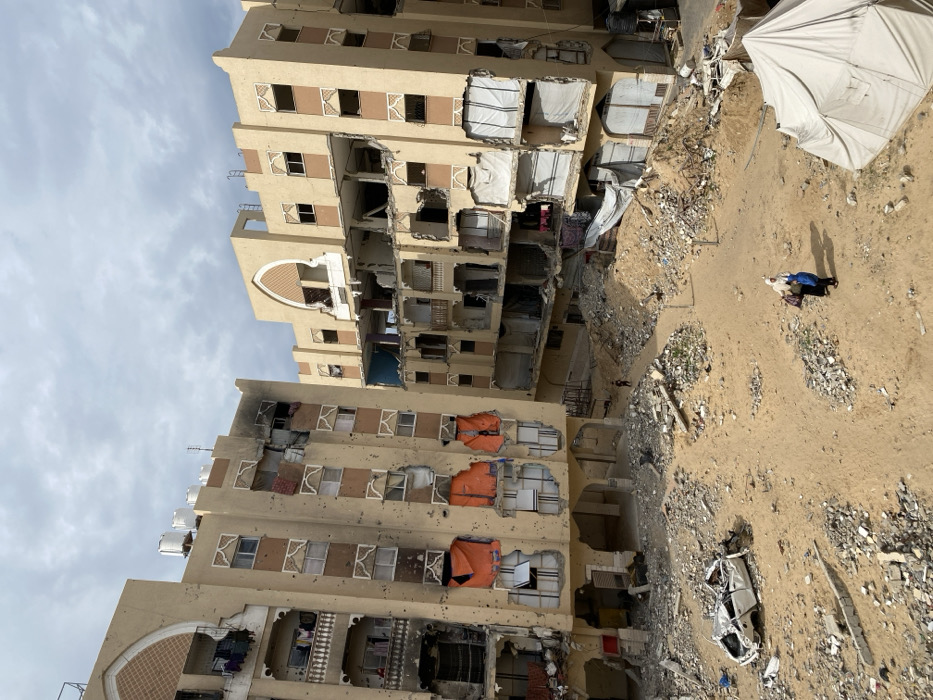The image depicts a scene of extensive structural damage in Khan Yunis, Palestinian Territory, under an overcast sky. Two multi-story residential buildings, constructed of light-colored concrete with arched architectural accents, dominate the frame. Both buildings exhibit significant destruction: the left structure features numerous impact holes across its facade, blackened areas indicative of fire damage, missing windowpanes, and several openings covered by orange tarps. Laundry is visible hanging on some surviving balconies. The right building shows major structural collapse on multiple floors, with entire sections of walls and interiors missing, revealing exposed rooms and debris, partially covered by white sheets or tarps.
The ground area between and in front of the buildings is a sandy, debris-strewn landscape. Piles of rubble, concrete fragments, and twisted metal are scattered across the sandy terrain. A heavily damaged white sedan, crushed and partially overturned, rests amidst the debris on the left side of the foreground. A dirt path winds through the sand and rubble. In the lower right foreground, two adults, one wearing a white hijab and dark clothing, the other in blue, walk on this path, carrying bags. Further back on the path, near the center, a smaller, solitary figure is visible. A white tent is partially seen in the lower right corner, nestled beside a mound of earth and debris.