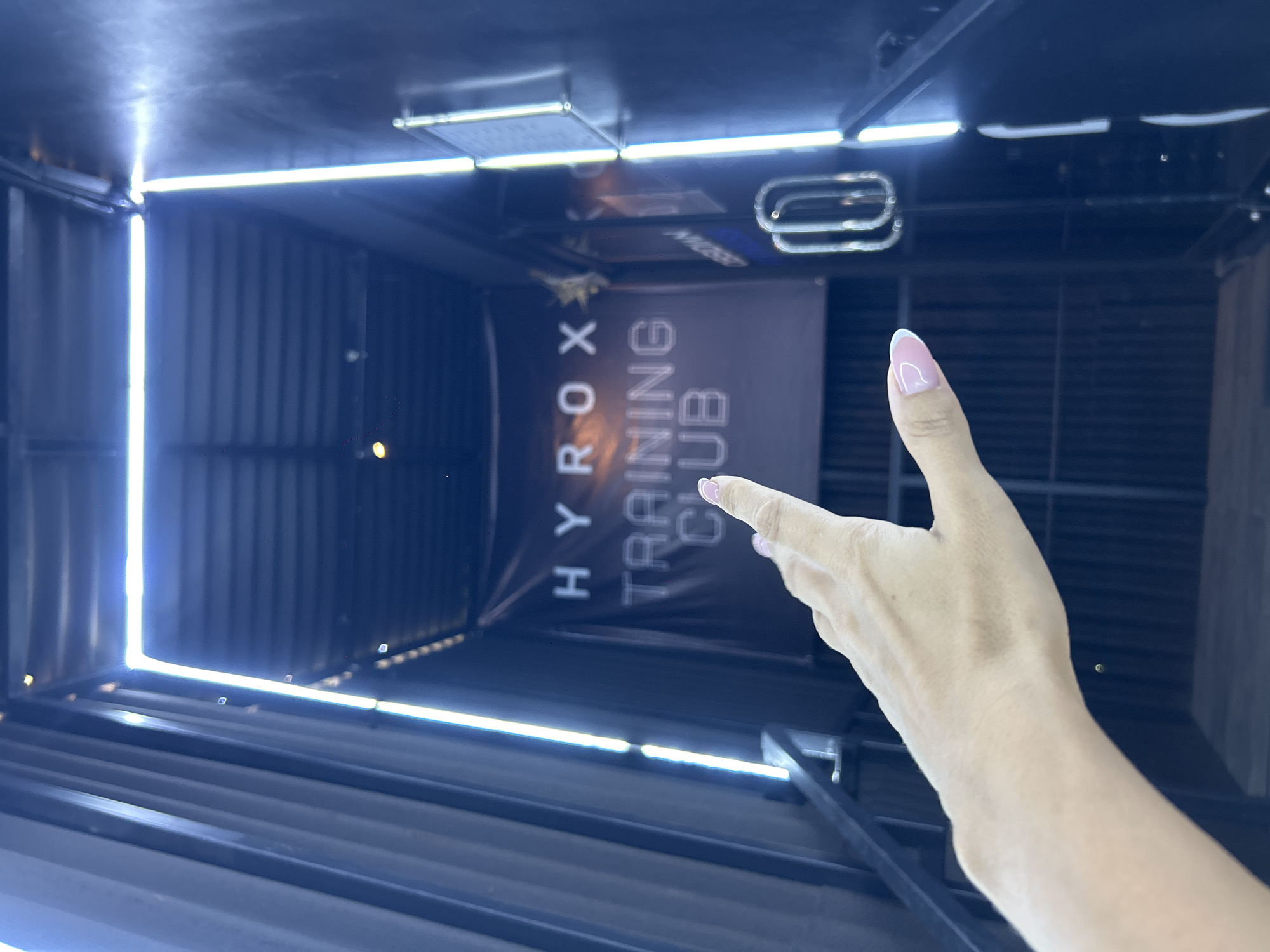 A close-up, first-person perspective shows a person's left hand, with a fingernail painted with a pink French manicure, pointing forward. The hand is positioned in the lower left quadrant of the frame, with the arm extending out of view. The background is a dimly lit, modern interior, featuring vertical corrugated metal walls and a dark ceiling with exposed beams. Bright white LED strip lights run along the edges of the ceiling and down the walls, illuminating a dark banner hanging in the center of the frame. The banner displays the words "HYROX TRAINING CLUB" in glowing white lettering, with "HYROX" in a sans-serif font and "TRAINING CLUB" in a more stylized, blocky font. To the right of the banner, a glimpse of a sign or poster with text is visible, along with a metal railing or structure. The overall impression is that of an entrance to a gym or training facility, possibly located in Mengwi, Indonesia, given the context.