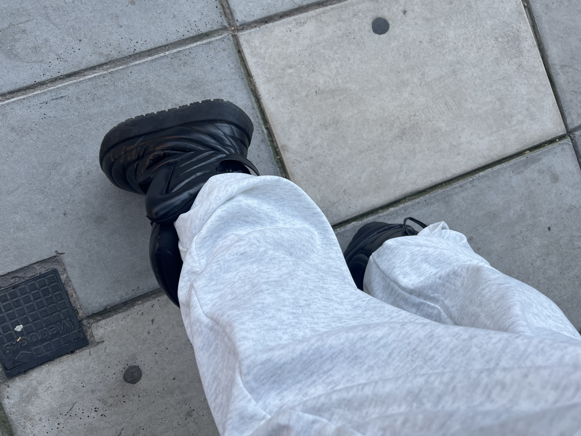 The image provides a close-up, high-angle perspective of a person's lower legs and feet on an outdoor concrete pavement. The individual is wearing light grey marl sweatpants, which appear soft and comfortable, gathered around the ankles. Their feet are clad in substantial black boots featuring a quilted or padded design, with a visible strap across the top of the left boot.
The setting is an urban environment, likely a sidewalk or public path, characterized by large, light grey concrete square tiles separated by dark grout lines. In the lower-left portion of the frame, a black rectangular utility cover with a grid pattern is visible. The word "MetroGas" is clearly inscribed vertically on this cover, confirming the location as Buenos Aires, Argentina, given that MetroGas is a prominent Argentine natural gas company.
Notable details on the pavement include a small, dark circular mark, possibly a stain, on one of the concrete tiles, and a tiny, metallic-looking circular object embedded in the concrete near the utility cover. The lighting is diffused and even, suggesting it is daytime, possibly under an overcast sky, as no strong shadows are cast. No specific activity is depicted beyond the casual stance of the person, implying they might be standing or walking. The overall color palette is dominated by greys and blacks.