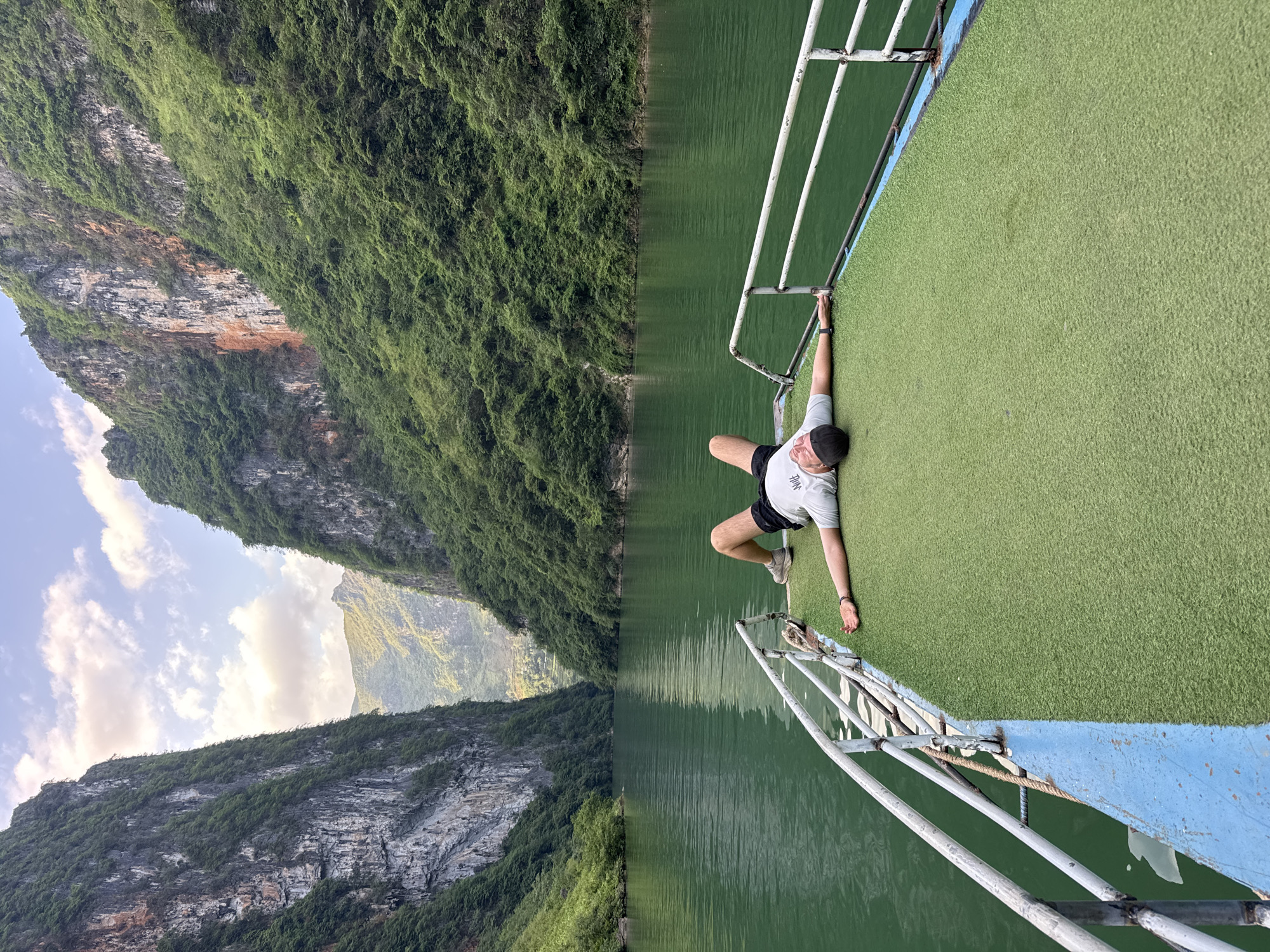 A person lies prone on the green AstroTurf deck of a boat, holding onto the metal railing with one hand. The boat appears to be traveling on a river or lake, with steep, forested mountains on the left side of the frame. The landscape suggests a location similar to the Đồng Văn Commune in Vietnam, characterized by rugged limestone karst formations covered in lush vegetation, and water. The boat's railing is made of white-painted metal, and the hull is painted light blue. The person is wearing a light-colored t-shirt, dark shorts, and sneakers. The water is dark green, and the sky is partly cloudy.