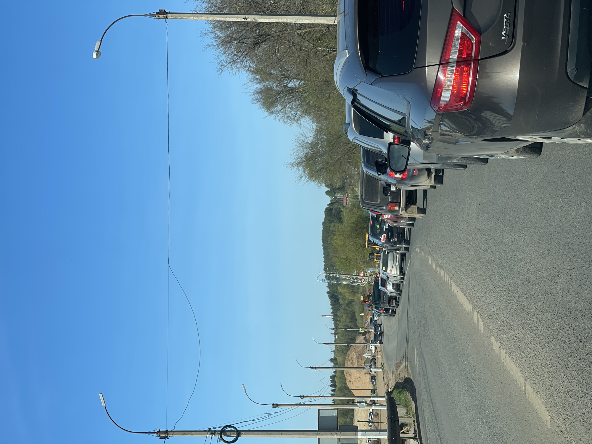 The image depicts a traffic jam on a paved road in Kabanikha, Russia, under a clear blue sky. Multiple vehicles, including a grey Lada Vesta, are lined up. Road construction is visible in the distance, potentially causing the congestion. Utility poles with streetlights line the left side of the road. Trees are present in the background.
