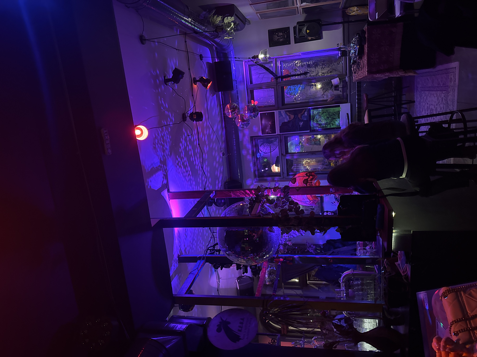 The image depicts the interior of K.E.B.A. bar in Sofia, Bulgaria, identifiable by a prominent circular sign with a rooster logo and "K.E.B.A." text on the left wall. The scene is illuminated predominantly by purple and magenta light sources, creating a vibrant, club-like atmosphere.
In the mid-ground, two individuals are seated with their backs mostly to the viewer, on black high-backed stools at a dark counter. Both have dark, long hair; one wears a visible white band around the waist of their dark attire.
The ceiling features exposed metallic ventilation ducts, multiple black track lighting fixtures, and a red "EXIT" sign. Numerous disco balls, including a large one centrally positioned within a dark structural frame and three smaller ones further right, project intricate light patterns onto the ceiling and walls.
The background comprises a wall with multiple window panes, adorned with string lights and various framed artworks or photographs of landscapes and abstract forms. To the far right, a table draped in a red, patterned cloth serves as a DJ station, equipped with audio mixers and associated electronics. A large black speaker stands next to it. Decorative elements include hanging plants, various glassware on shelves behind the counter, and an antique-style red lampshade. In the immediate bottom-left foreground, a white quilted bag with a gold chain strap rests on a surface.