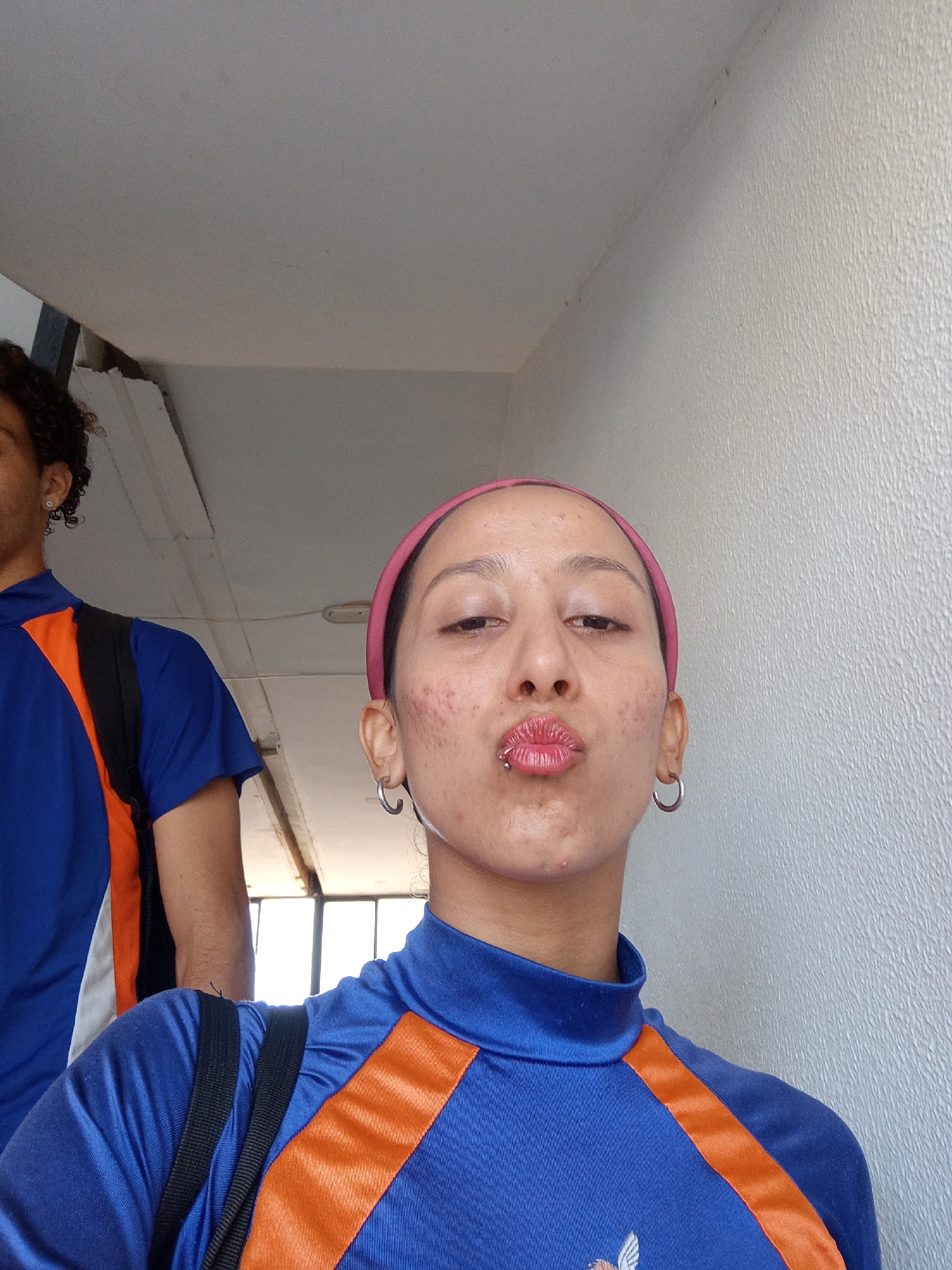 A medium shot depicts a young woman with light brown skin, dark eyes, and a pink headband, making a kissing face toward the camera. She wears a blue and orange athletic top and silver hoop earrings and has a lip piercing. A man with curly dark hair, also in a matching athletic shirt, is partially visible to her left and slightly behind her. Both individuals wear black backpack straps, and they appear to be standing on a stairway with a white wall and a window in the background. The photograph may have been taken indoors, possibly in a building in Porlamar, Venezuela.