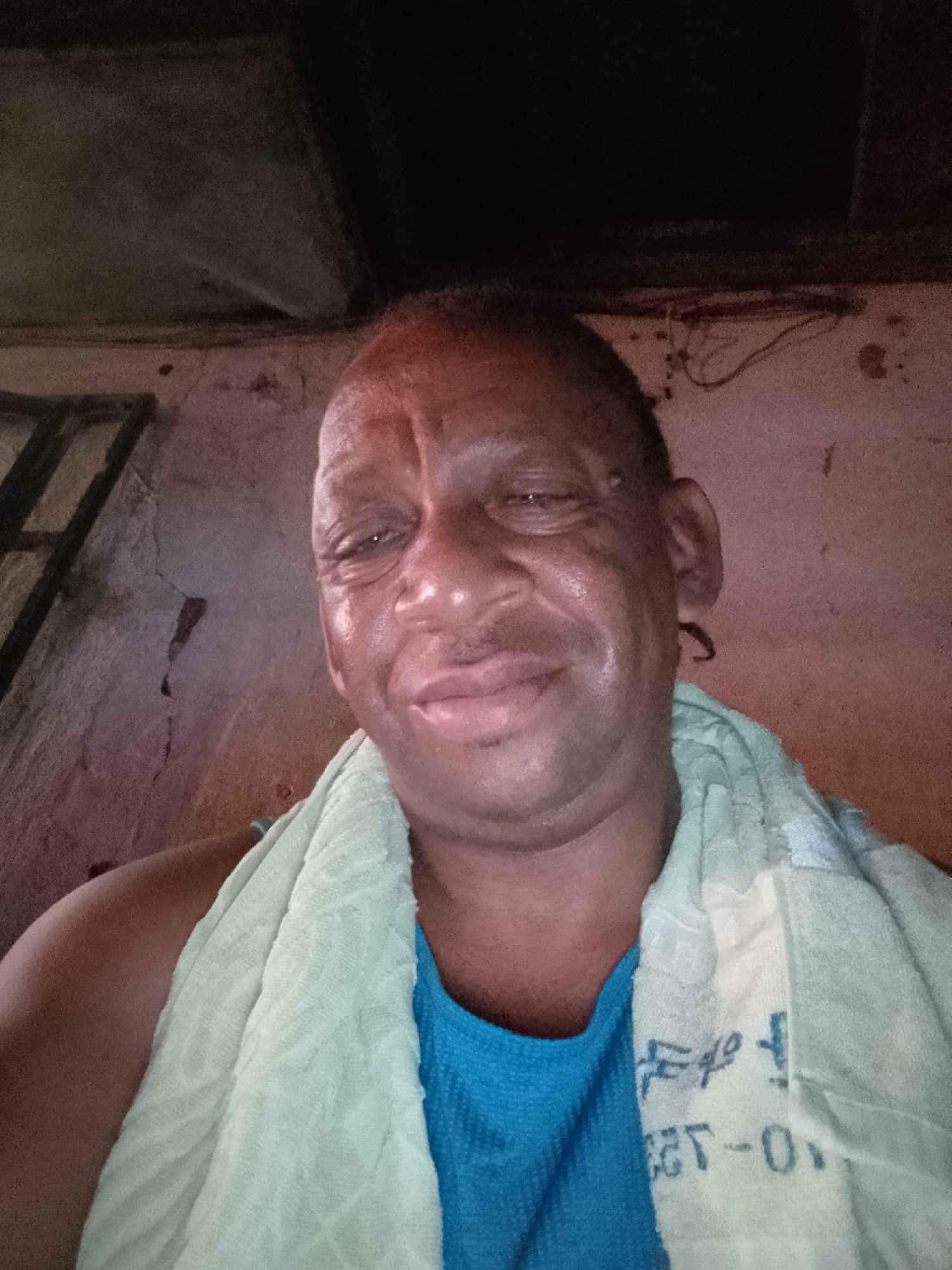 A selfie of a man with dark skin wearing a blue shirt and a light green towel around his neck. The location is possibly indoors based on the unfinished wall and ceiling. The city of Sabon Tasha, Nigeria, is not verifiable from this image.