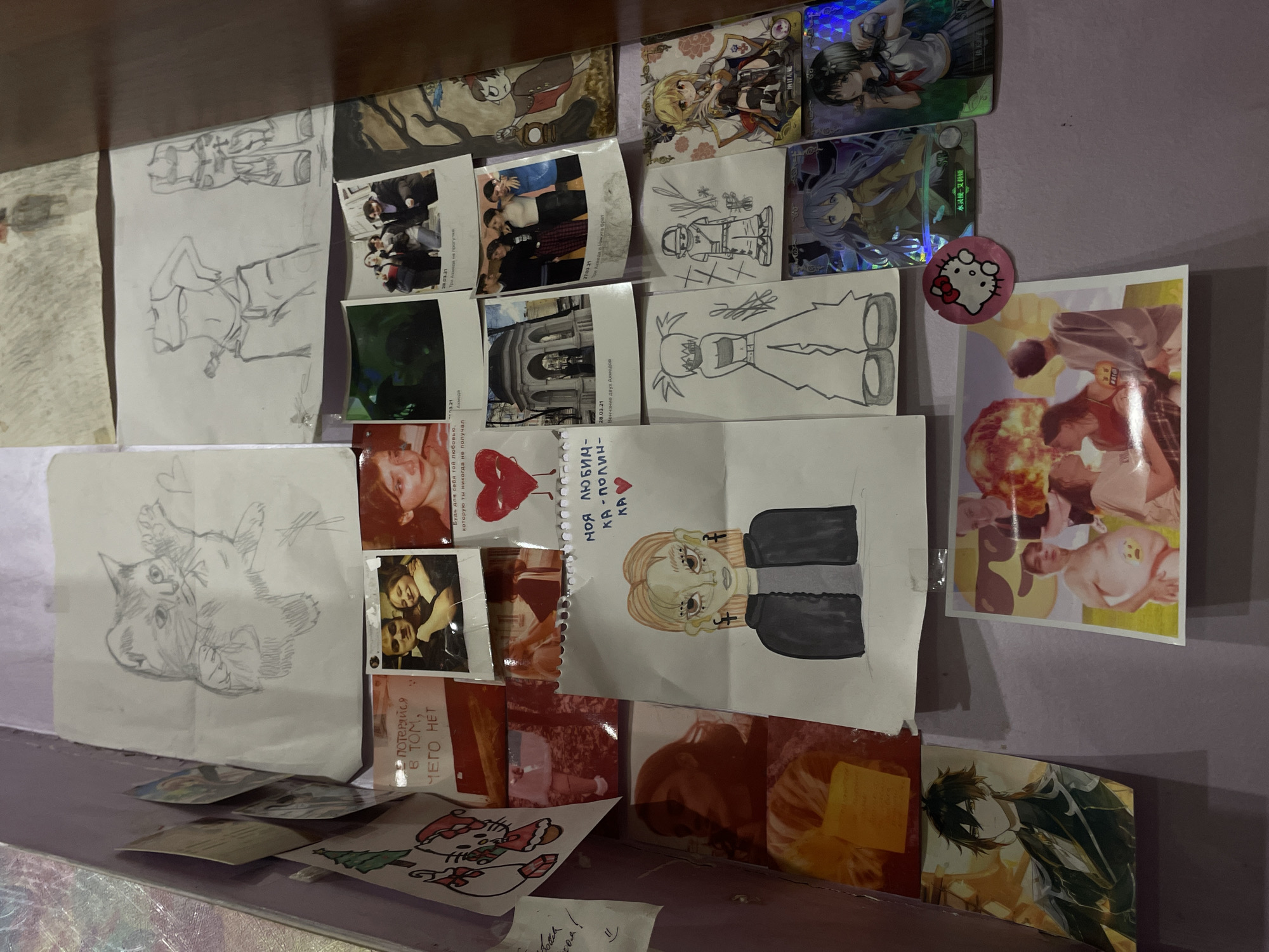 The image displays a wall covered with various items, including drawings, photographs, and what appear to be trading cards. The overall impression is one of personal display, possibly a bedroom or a creative space. The items are arranged in a collage-like fashion.
On the left side, there is a drawing of a cat. Below this are several photographs. One is a Polaroid-style photo of a couple embracing. Another shows a person's face in profile, appearing distressed. There are also images with text overlays.
In the center, a drawing of a person with blonde hair and a dark jacket is prominent. Above this drawing, there are more photographs, including one of a group of people outside a building and another of a couple. A drawing of a heart with a face is also visible.
To the right, there are drawings of figures in casual attire, and several trading cards featuring anime-style characters. A circular Hello Kitty sticker is also present.
The background wall is a solid, muted color, possibly purple or lavender. The lighting suggests an indoor environment, possibly with artificial light sources contributing to the overall ambiance. There is no visible indication of the specific location within the city of ЖК «Борисоглебское», Russia. The image focuses solely on the decorated wall.