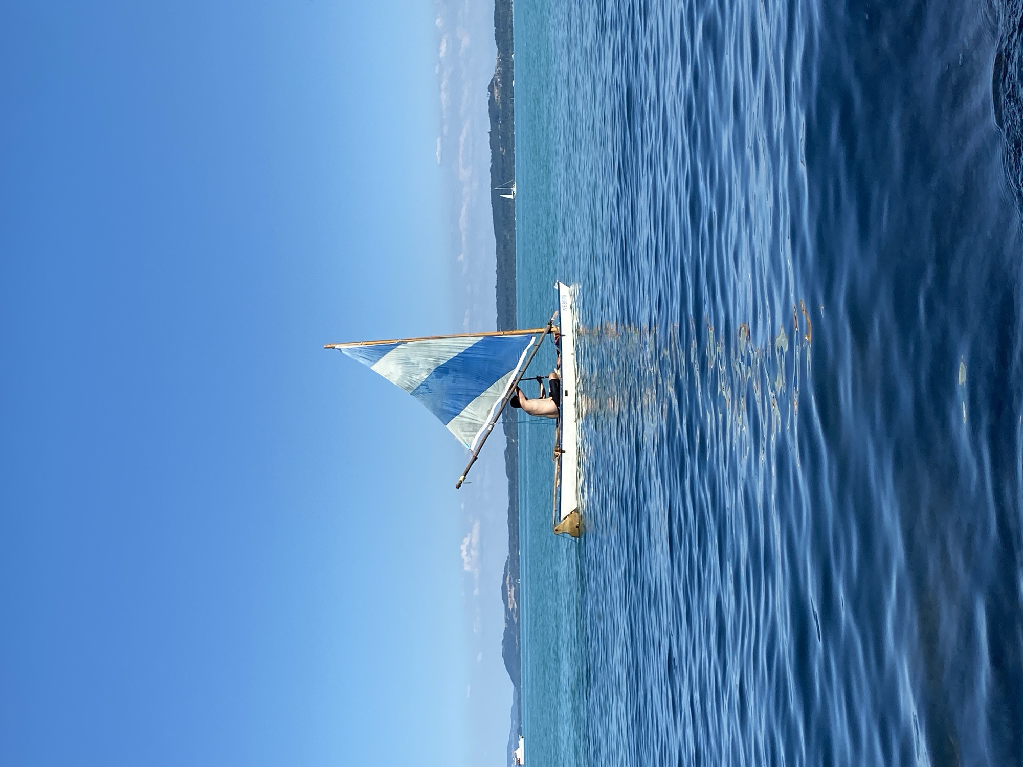 A person is sailing a small sailboat on a calm, blue body of water under a clear sky. The sailboat has a white hull and a triangular sail with blue and white stripes. The person, who appears to be male and is shirtless, is seated in the boat and steering. In the background, there is a distant landmass with rolling hills and a few other sailboats. The water is slightly rippled. The image is taken from a low angle, placing the viewer on the water's surface. The location of Cortaillod, Switzerland is not evident from the image.
