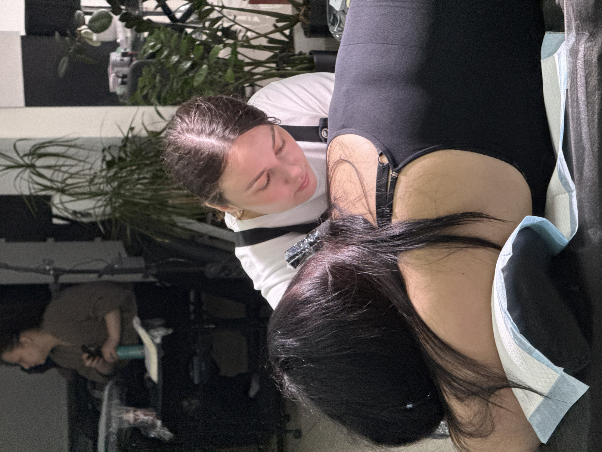 A person in a white top is examining the side of another person in black clothing who is lying down. The person in black has dark hair and is positioned on a surface covered with a disposable absorbent pad. In the background, there is a plant and a partial view of another person. The location and city are not definitively discernible from the image.