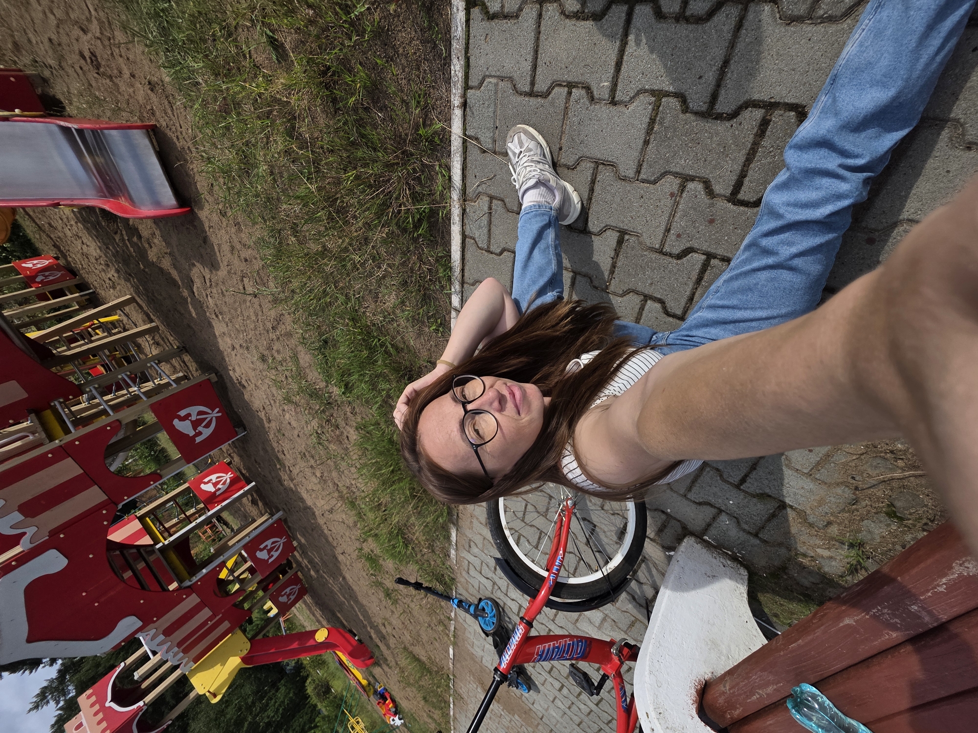 A young woman with long brown hair and glasses sits on a paved area next to a red bicycle and a scooter. She is wearing a striped tank top and blue jeans. Behind her is a playground with a large red and yellow structure, slides, and a sandy area. In the foreground, a brown wooden bench is partially visible, with a blue plastic water bottle on it. The scene appears to be outdoors on a sunny day, possibly in a park or recreational area in Chaikovsky, Russia.