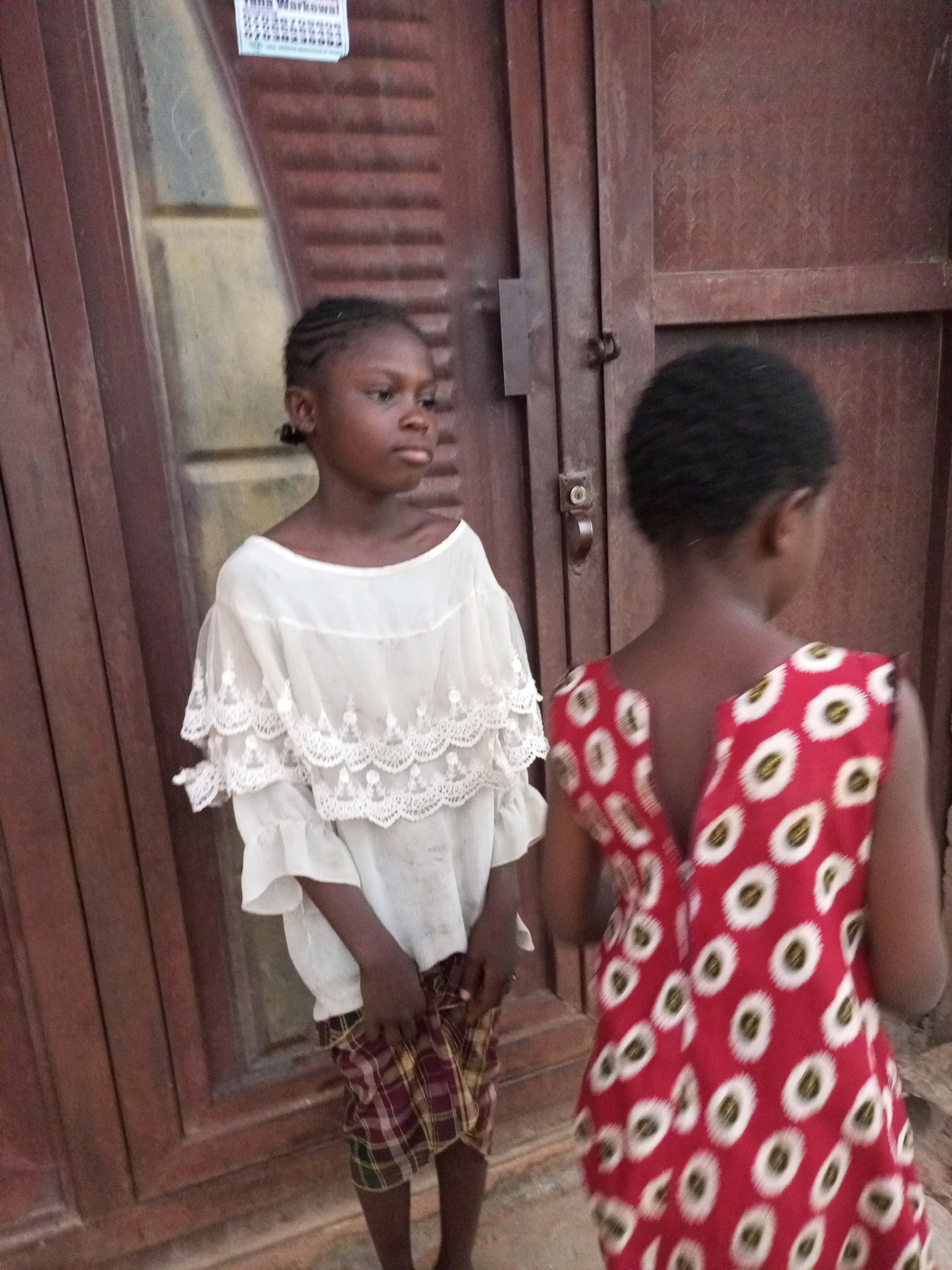 In Tempure, Nigeria, two young girls stand in front of a wooden door. The girl on the left wears a white, tiered lace top and a plaid skirt, her braided hair pulled back. She faces right, looking away from the camera. The girl on the right, facing away from the camera, wears a red dress with a white circular pattern. She has dark, curly hair. The wooden door appears aged, with a dark, reddish-brown hue. A notice is affixed to the upper left portion of the door. The ground beneath them is composed of rough, gritty material.