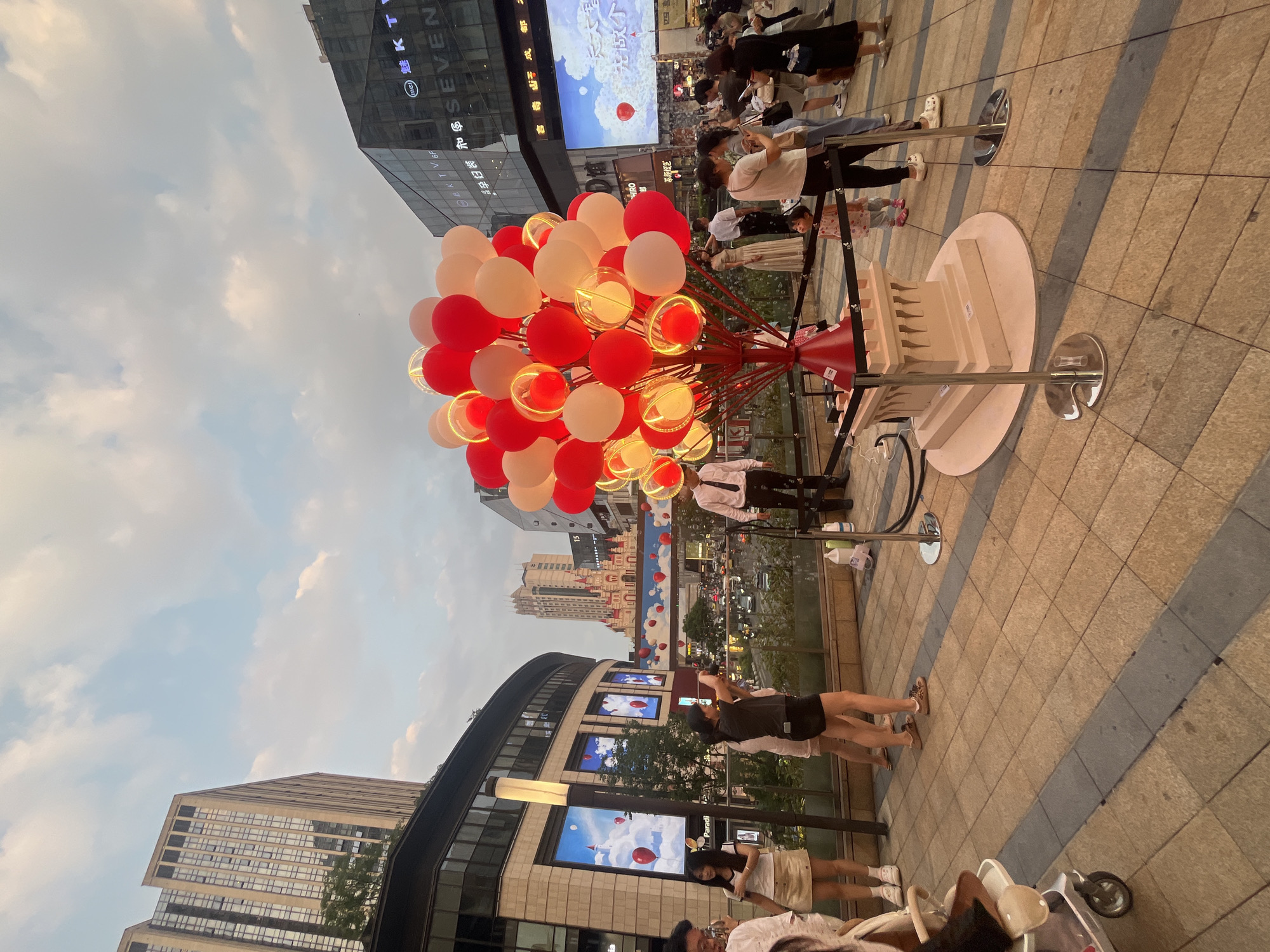 The image depicts an elevated outdoor urban plaza or pedestrian area in Yuzhong District, China, at dusk. The sky above shows a soft pale blue with scattered white clouds, indicating the transition from late afternoon to early evening, with artificial lights already active.
In the foreground, slightly right of center, stands a large, illuminated art installation resembling a balloon tree. It features numerous red and white/cream balloons, some of which are clear spheres with internal warm-toned lights, arranged on a branched red structure emerging from a light-colored, stylized castle turret base. This installation is cordoned off by black ropes supported by silver stanchions.
Numerous people populate the plaza. In the left foreground, a person's head and shoulder are visible, with a baby stroller partially seen. Next to a modern lamppost, a young woman in a white top and beige skirt stands, looking left. Another woman in a black top and dark shorts is actively taking a picture or video with her smartphone, facing towards the right. Behind the balloon sculpture on the left, a man in a white shirt and dark tie stands. On the right side of the sculpture, a small child in a patterned top looks intently at the balloons, accompanied by an adult woman in a light top and dark pants. Various other individuals are visible in the mid-ground, walking, standing, and observing the scene.
The background features several modern buildings. On the left, a tall, light-colored high-rise building with numerous windows rises above a multi-story commercial structure. This lower structure displays multiple vertical LED screens showing animated red balloons against a blue sky. In the mid-ground, a unique, multi-turreted building with red roofs, resembling a stylized castle, stands behind a dark-glassed commercial building on the right. This right-hand building features large signage including "KTV" and "SEVEN" and hosts a very large LED screen displaying an advertisement, also featuring balloons. Below the pedestrian level, a busy street with vehicle traffic is visible, separated by a dark railing. The ground of the plaza is paved with a pattern of light-brown and grey square and rectangular tiles.