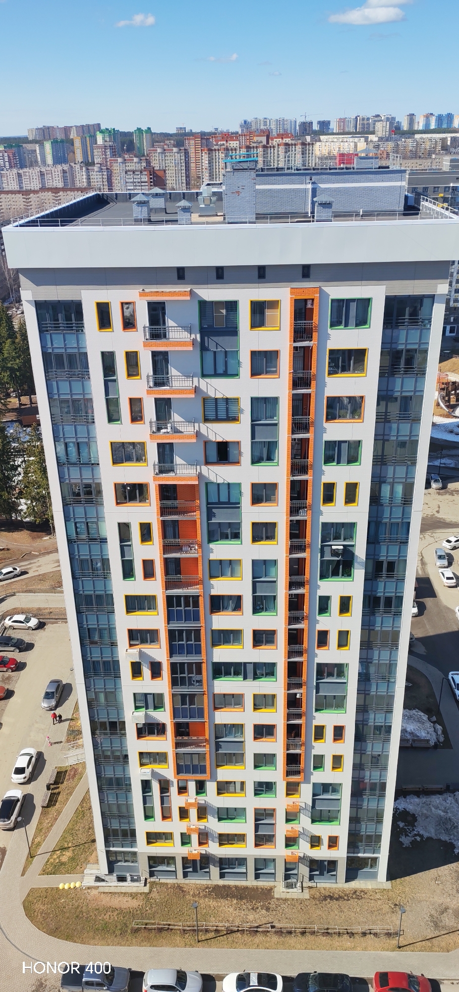 The image shows a tall, modern residential building with a distinct architectural design in Izhevsk, Russia. The building has a gray and white exterior with colorful window frames in orange, yellow, and green. It has a central section with balconies and smaller windows, flanked by sections with large glass windows. The top of the building is flat and gray. In the background, there is a view of other residential buildings and city structures under a clear, blue sky. The foreground shows a parking area with several cars, a walking path, and some green space. Two people are visible walking on the path. Patches of snow are visible near the building.