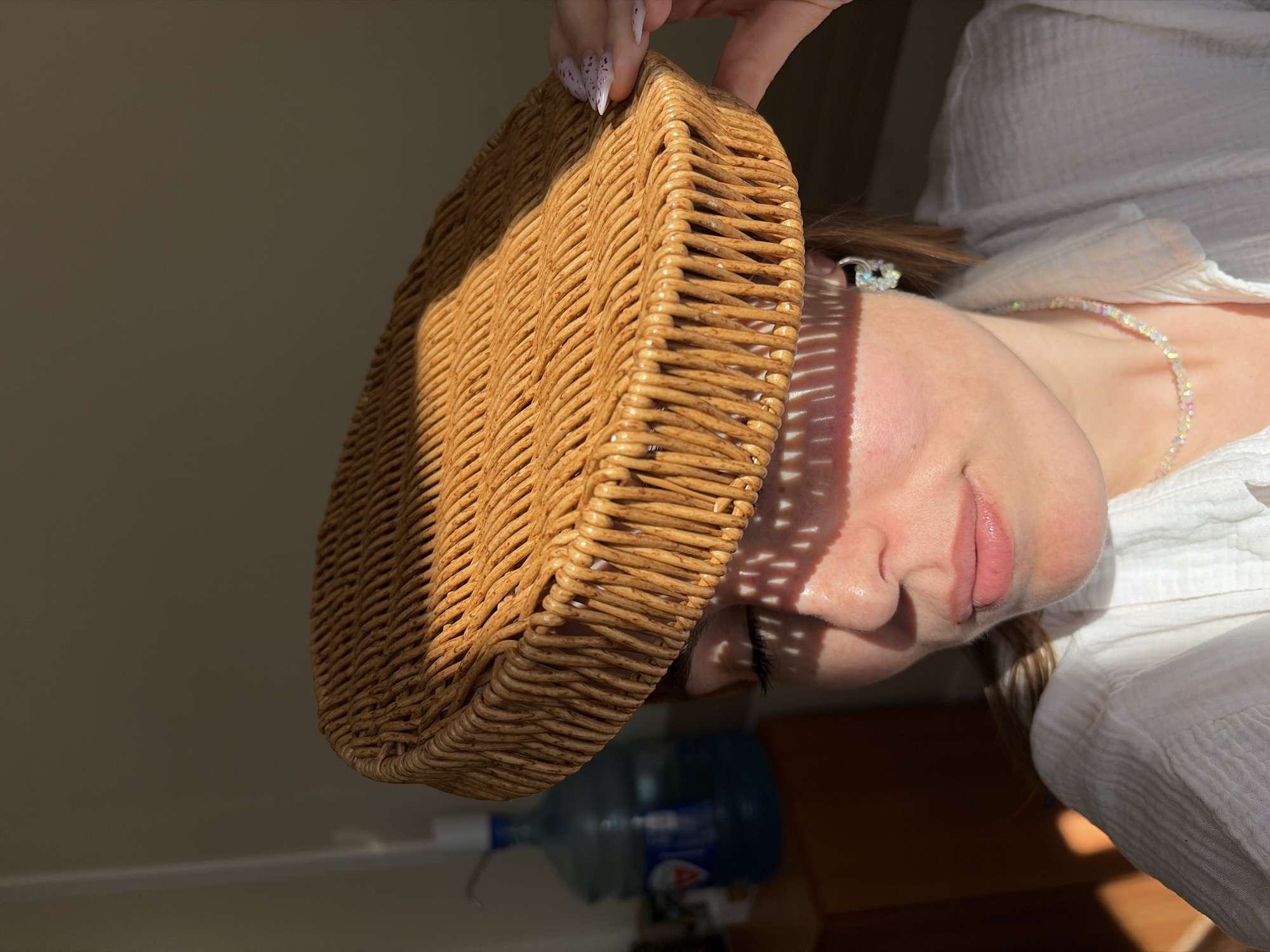 A medium shot depicts a woman holding a round, woven basket over her head. The basket casts a patterned shadow on her face. She is wearing a white, collared shirt and a beaded necklace. Her nails are decorated with white designs. A large water bottle is visible in the background. The location and whether it is in the city of Izhevsk, Russia, is not determinable from the given information.