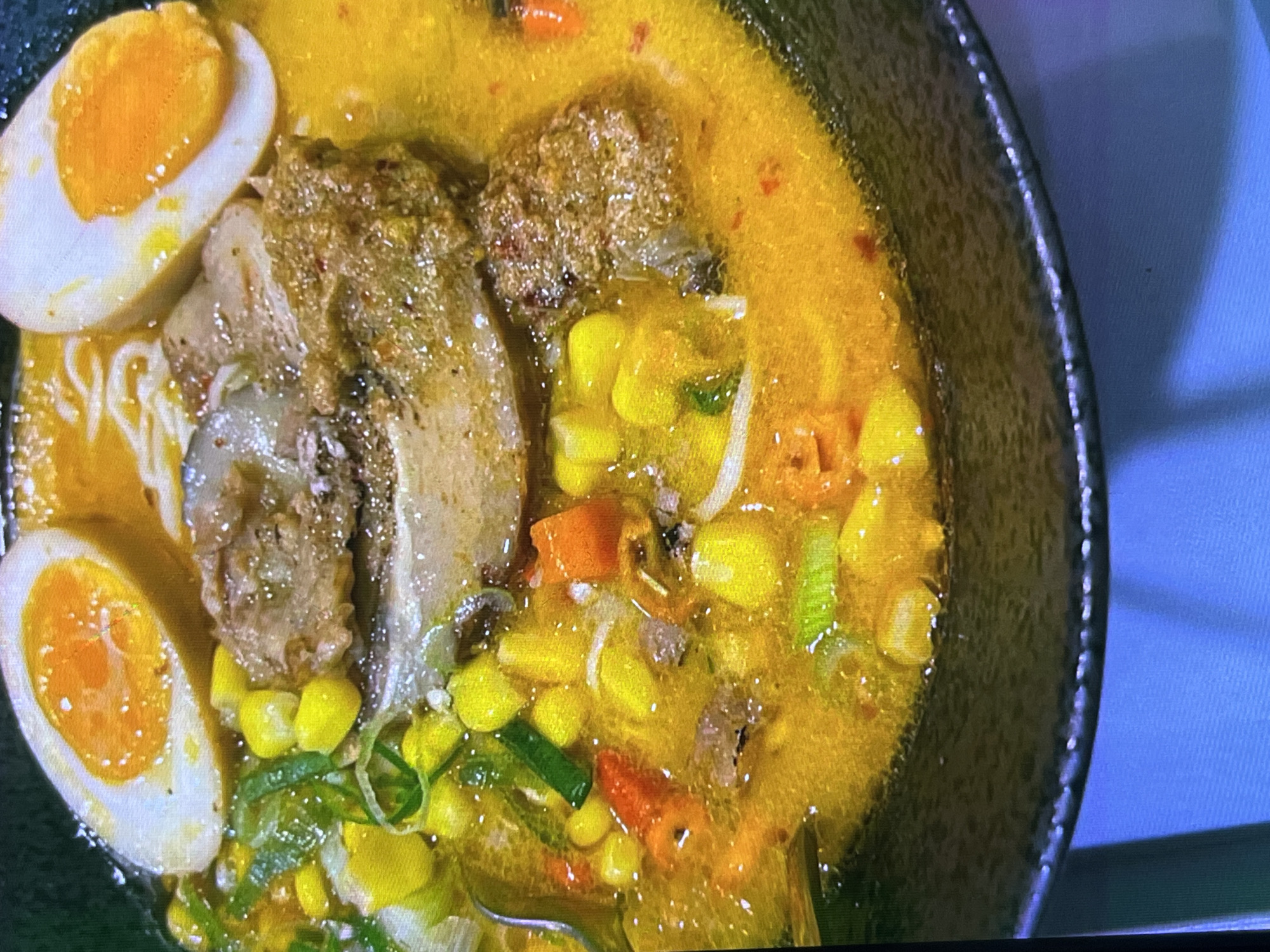 This is a close-up shot of a bowl of ramen soup, likely a hearty and flavorful variety given the ingredients. The soup base is a vibrant, golden-yellow broth, suggesting a rich broth, possibly miso or curry-based. Submerged within the broth are several key components.
Prominently featured are two halves of a soft-boiled egg, each with a perfectly cooked, bright orange yolk and firm white. There is also a substantial piece of braised pork, its texture appearing tender and slightly marbled. Scattered throughout the soup are bright yellow kernels of corn, small pieces of orange carrot, and chopped green scallions, adding color and texture. Thin white noodles are visible beneath the surface of the broth, indicating this is a noodle soup dish. The bowl itself is dark and appears to be made of ceramic, with a slightly textured exterior.
The lighting suggests an indoor setting, possibly a restaurant or home kitchen, and there are no overt cues about the time of day or weather. The overall impression is one of a delicious and satisfying meal. The image does not contain any visible text. Given the location context of Cadek, Indonesia, this ramen could be a local adaptation or a popular international dish being enjoyed there.