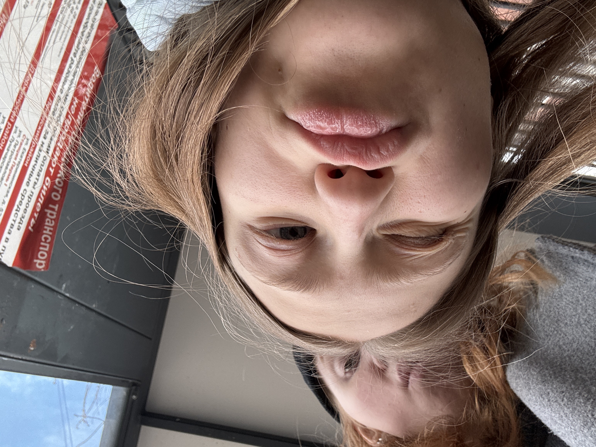 A selfie angled upward features a young woman with blonde hair and one eye closed. A sign in the background indicates the location is in Perm, Russia, specifically near the Yekaterininskaya street, Perm I station, Petropavlovskaya street, and Lunacharsky street. The sign states "Drivers do not sell tickets" in Russian and that you should pay for travel independently.