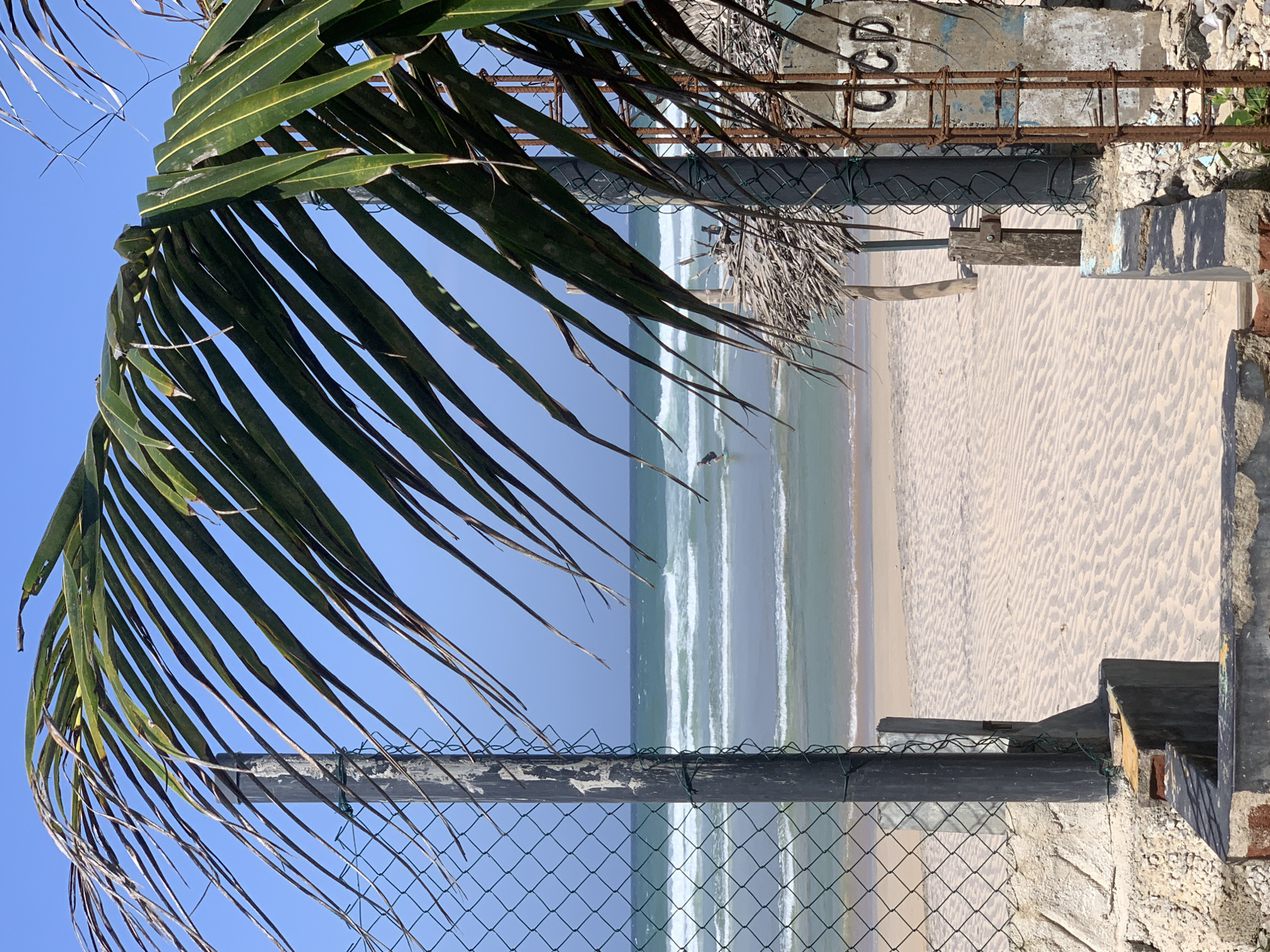 A scene unfolds at a beach in Ahangama, Sri Lanka, viewed through a gap in a wire fence and framed by the fronds of a palm tree. The foreground is dominated by a rough, concrete and brick structure forming part of a broken fence. Beyond this, a wide expanse of sand stretches to meet the ocean. The sand exhibits a rippled texture, suggesting the ebb and flow of the tide or wind.
The ocean is a vibrant turquoise, with distinct layers of waves rolling towards the shore. The waves are moderate, breaking with white crests. A lone figure, appearing to be a surfer, is visible in the water, positioned further out from the shore.
To the right of the frame, a rustic thatched structure, possibly a small hut or shelter, stands partially obscured by the fence and palm leaves. The background is a clear, bright blue sky. The overall impression is one of a tropical beach setting, with signs of coastal development or decay.