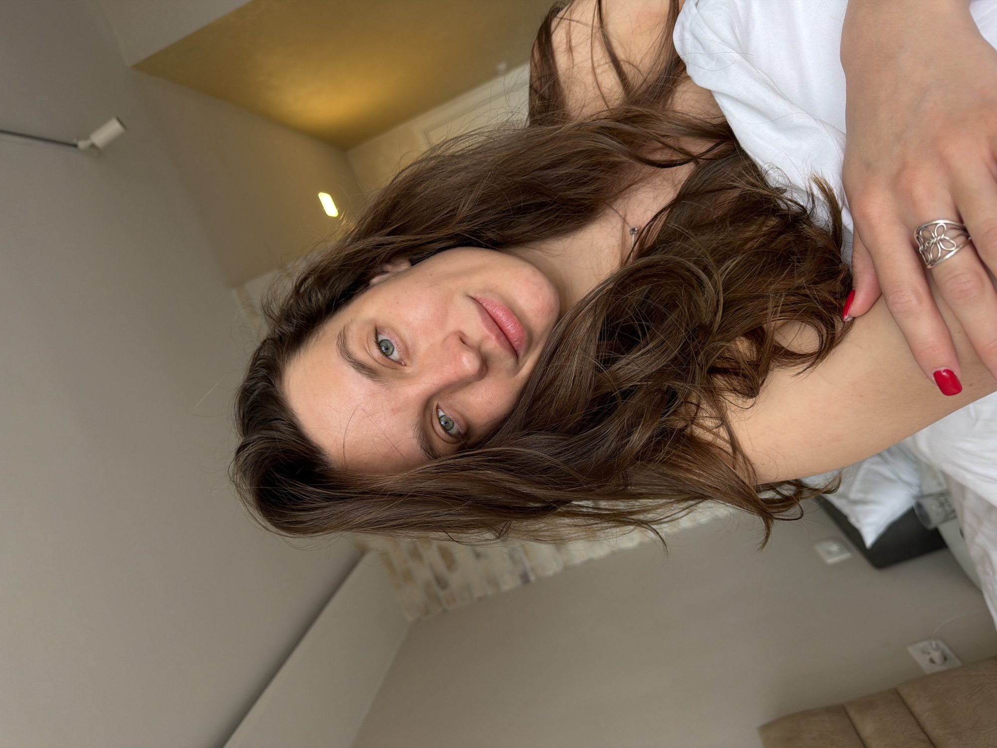 A young adult woman with fair skin and long, brown hair is lying down, possibly in bed, in a brightly lit interior. She has light-colored eyes and a slightly pink complexion. Her right hand, adorned with a silver flower-shaped ring and red nail polish, is visible. She's under white sheets. The background includes a wall and a ceiling with light fixtures. There is no specific evidence in the image to confirm that it was taken in Budva, Montenegro.