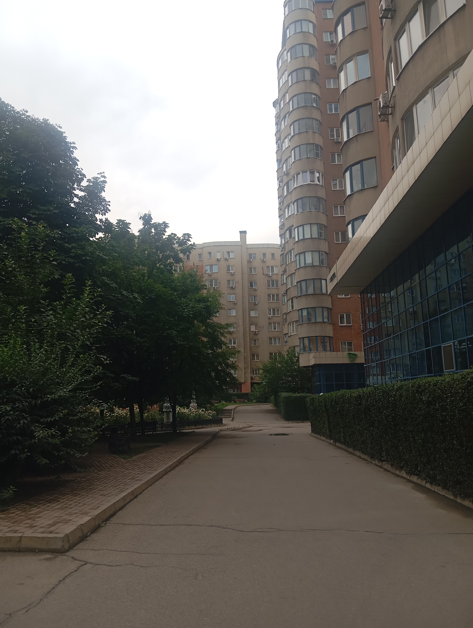 The image depicts a street scene in a city, potentially Almaty, Kazakhstan, with apartment buildings, trees, and a paved walkway. On the right side of the frame, a modern building with a glass facade and a prominent overhang dominates the foreground. Adjacent to it, a tall, curved apartment building with many windows and balconies rises vertically. To the left, a dense cluster of trees with dark green foliage lines the walkway, partially obscuring another apartment building in the background, which appears older and lighter in color. The walkway itself is paved with bricks and bordered by a concrete curb. The street in front is asphalt with visible cracks. The sky is overcast and a pale gray. The scene suggests an urban residential area, possibly a courtyard or a section of a larger complex. There are no people visible in the image.