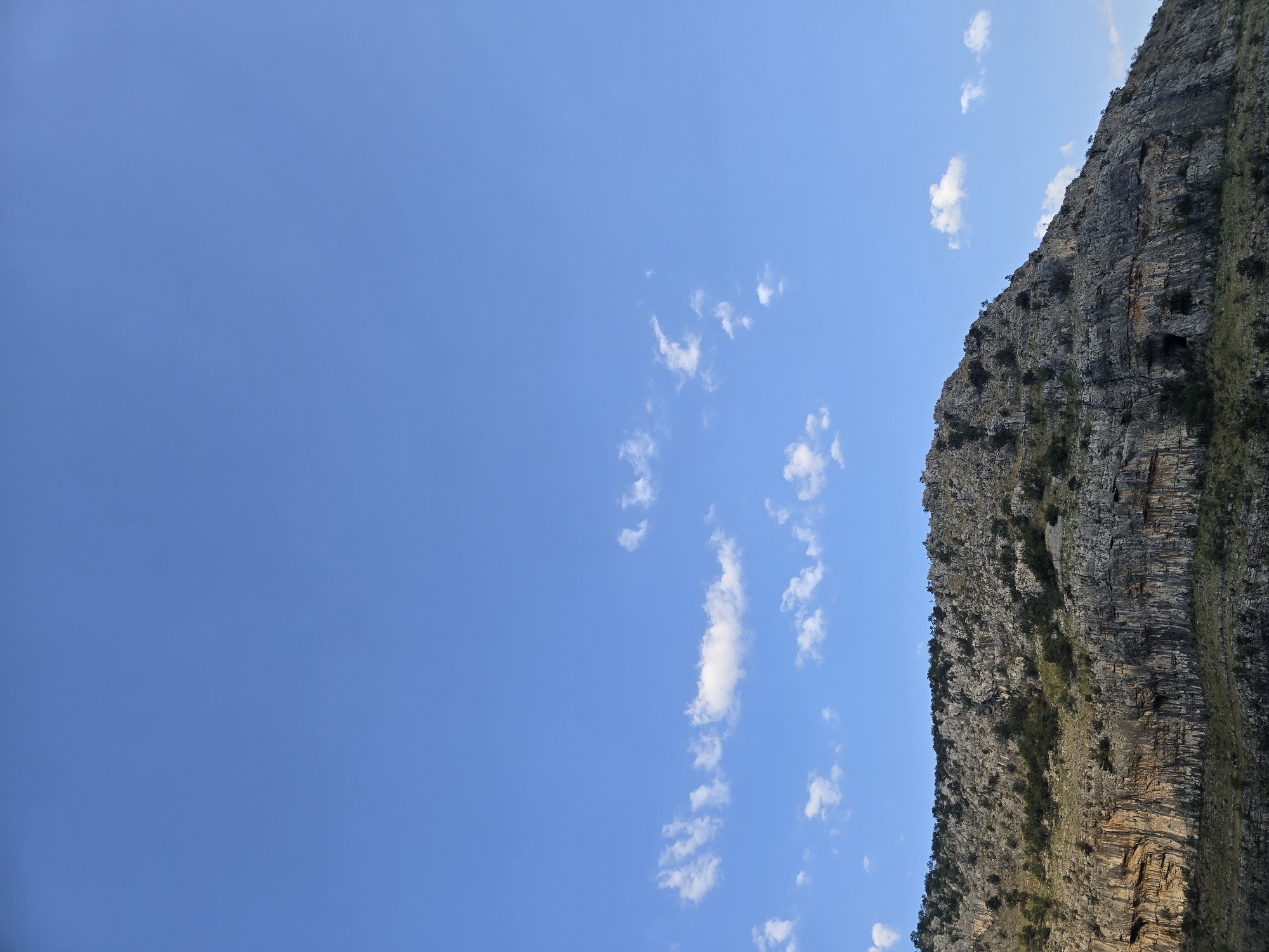 A wide shot captures a rugged, rocky mountain against a vast, clear blue sky. Sparse white clouds are scattered across the upper portion of the sky. The mountain itself is covered in a dense layer of gray rock, with visible layers and textures. Small green trees and shrubs are interspersed across the mountain's surface, particularly along its crest and lower slopes. The lower part of the mountain shows evidence of erosion and has a stratified appearance, with darker rock formations and some lighter, weathered sections. There are no people, animals, or man-made structures visible in the image. The lighting suggests it is daytime, with sunlight illuminating the mountain face.