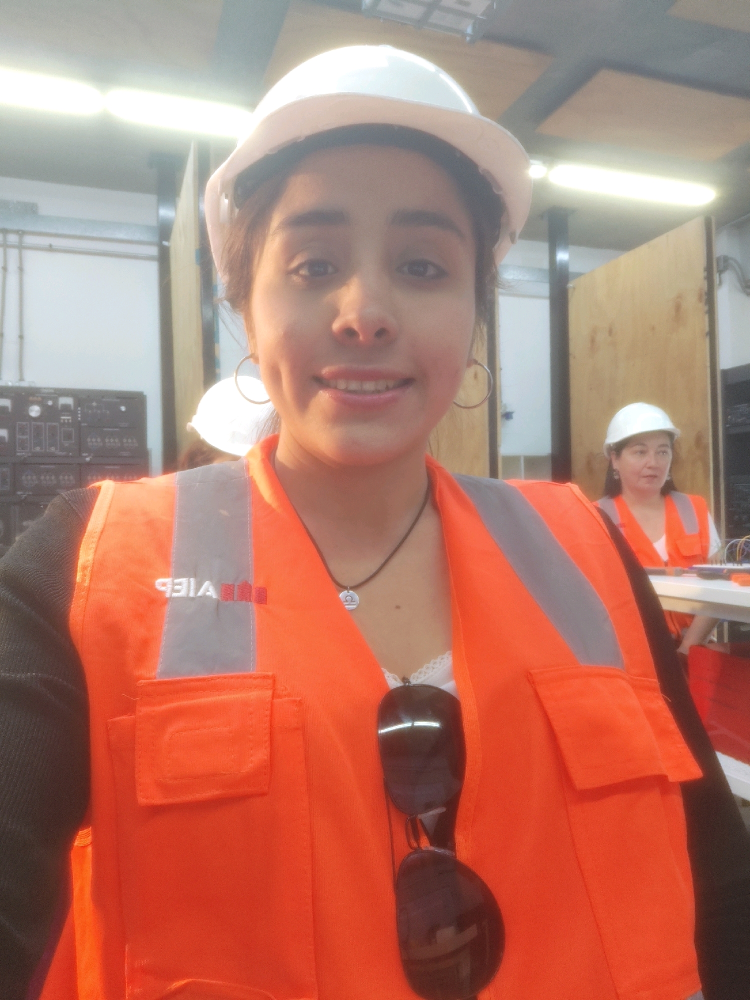 The image is a selfie taken by a young woman wearing a white hard hat and an orange safety vest over a white garment with lace trim. She has brown hair, brown eyes, and is wearing large hoop earrings and a necklace with a zodiac sign pendant. Dark sunglasses are hanging from the front of her vest. To her right, another woman in a similar hard hat and orange safety vest is visible, looking to her left. They appear to be indoors in a workshop or laboratory setting with wooden walls and overhead lighting. In the background on the left, there is a rack of electronic equipment. The overall impression is of a vocational or educational environment.