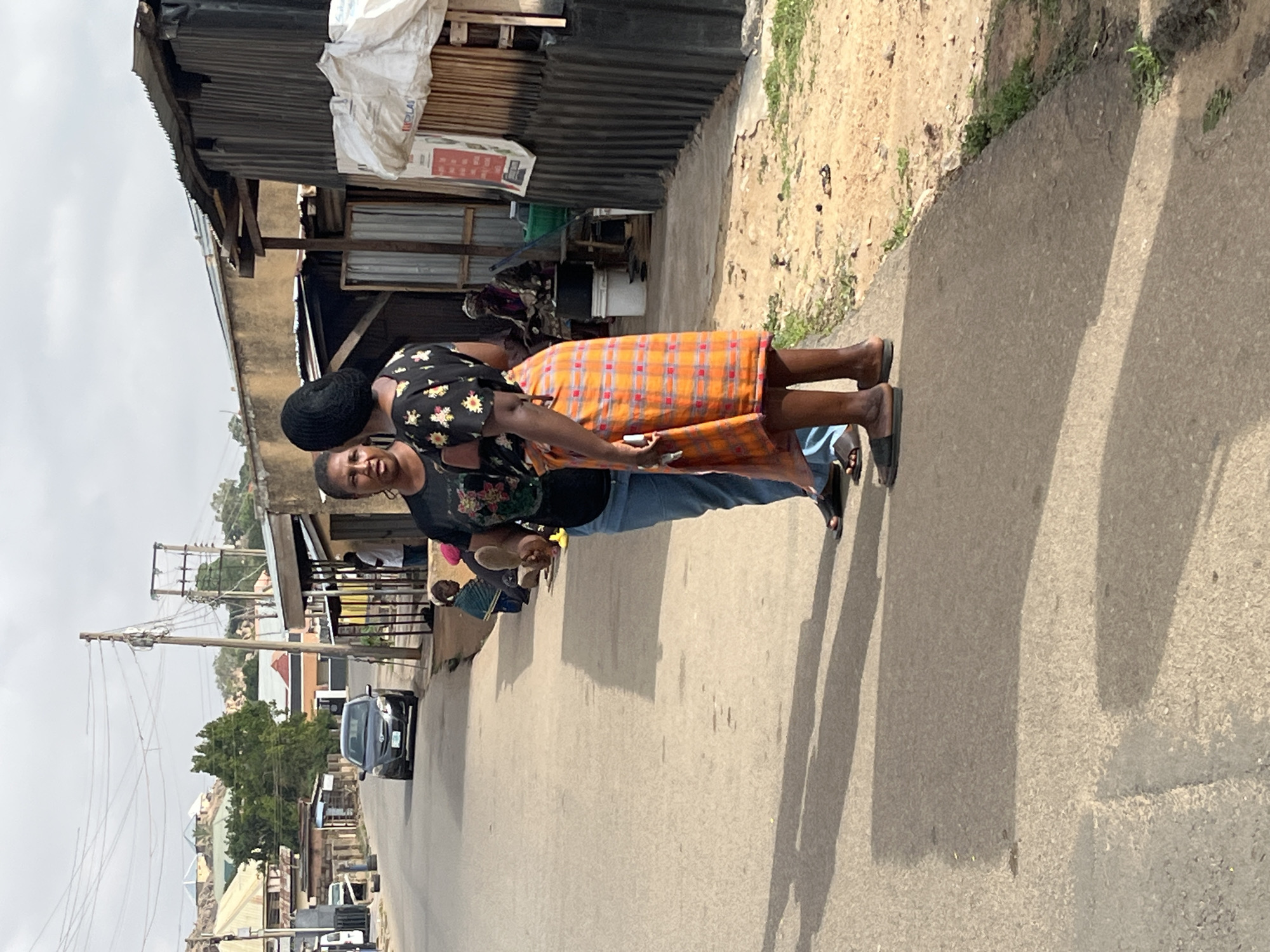 Two women stand on a concrete sidewalk next to a street in Jos, Nigeria. The woman closer to the camera wears a black t-shirt with floral print, blue jeans, and sandals, and has short hair and a black head covering. The other woman wears a black top with floral print and a plaid orange and white patterned skirt, also with sandals. Their hands are touching or holding each other. They stand in front of buildings with corrugated metal siding and other structures typical of a residential or commercial area. The street is paved and leads to more buildings, utility poles, and vehicles in the distance. The sky is overcast.