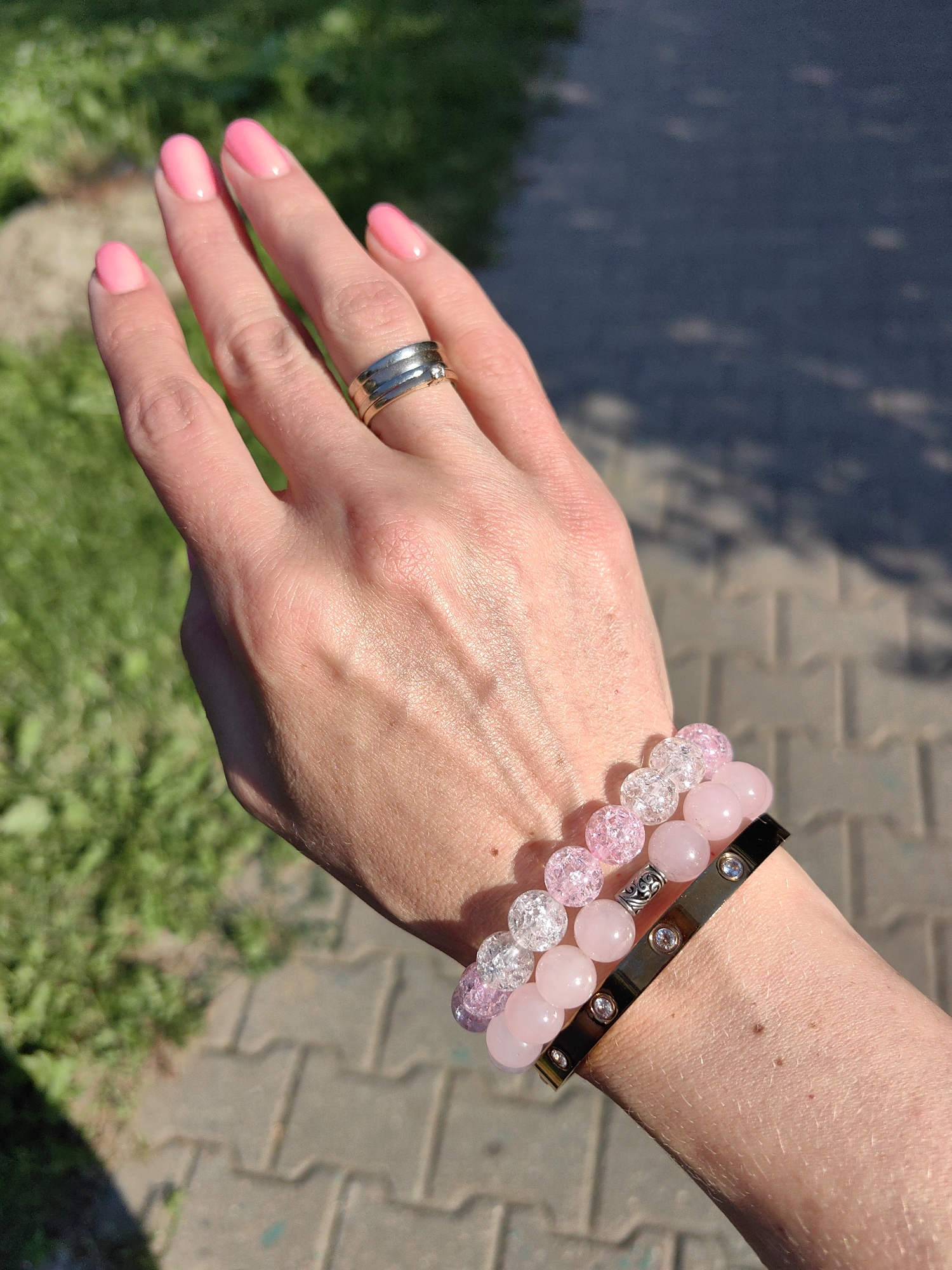 A close-up, eye-level shot captures a woman's left hand adorned with a silver ring and two beaded bracelets. The ring, worn on the ring finger, is a stack of three silver bands, one of which features a small diamond. The bracelets, stacked on the wrist, consist of pink and clear beads, with one bracelet being a silver cuff adorned with small, embedded clear stones. The hand is positioned against a blurred backdrop of green grass and a paved pathway. The lighting suggests it is daytime, with sunlight casting shadows. The scene appears to be outdoors, possibly in a park or a residential area. There is no discernible indication of the location being in Minsk, Belarus.