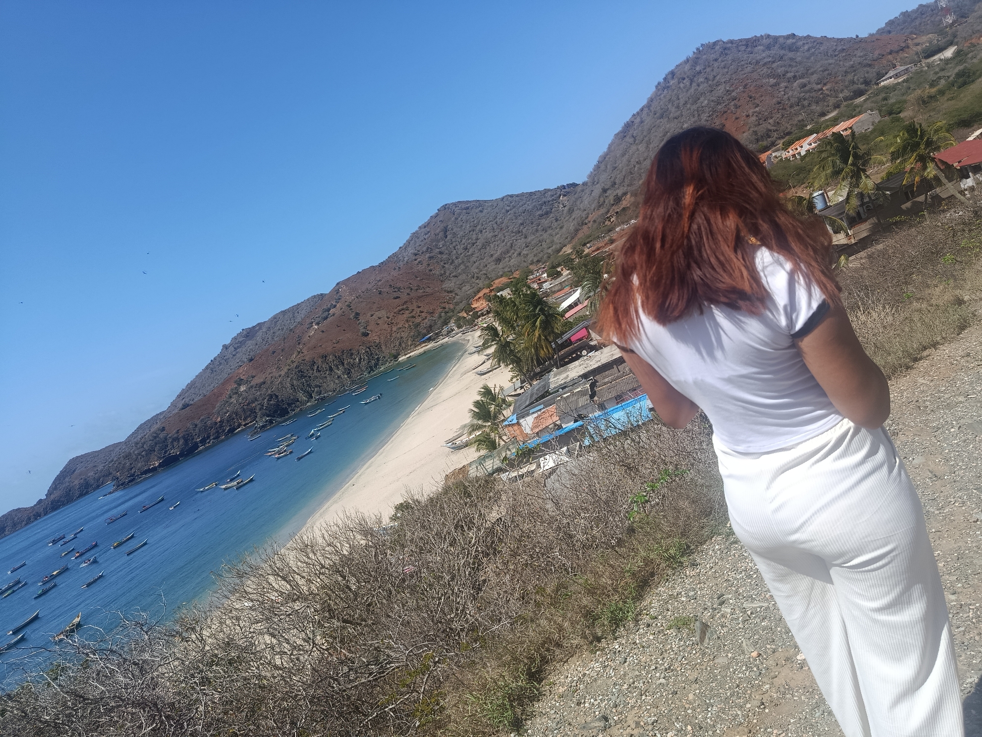 The image shows a woman from behind, wearing a white t-shirt and white ribbed pants, standing on a gravel path overlooking a scenic bay. The bay features calm blue water with numerous small boats anchored offshore. A white sandy beach lines the bay, backed by a small village with buildings and palm trees. Steep, arid, brown hills rise behind the village, extending towards a clear blue sky. Several dark specks, possibly birds, are visible in the sky. The overall scene depicts a coastal landscape with human habitation. The location is identified as the bay of Manzanillo, Venezuela.