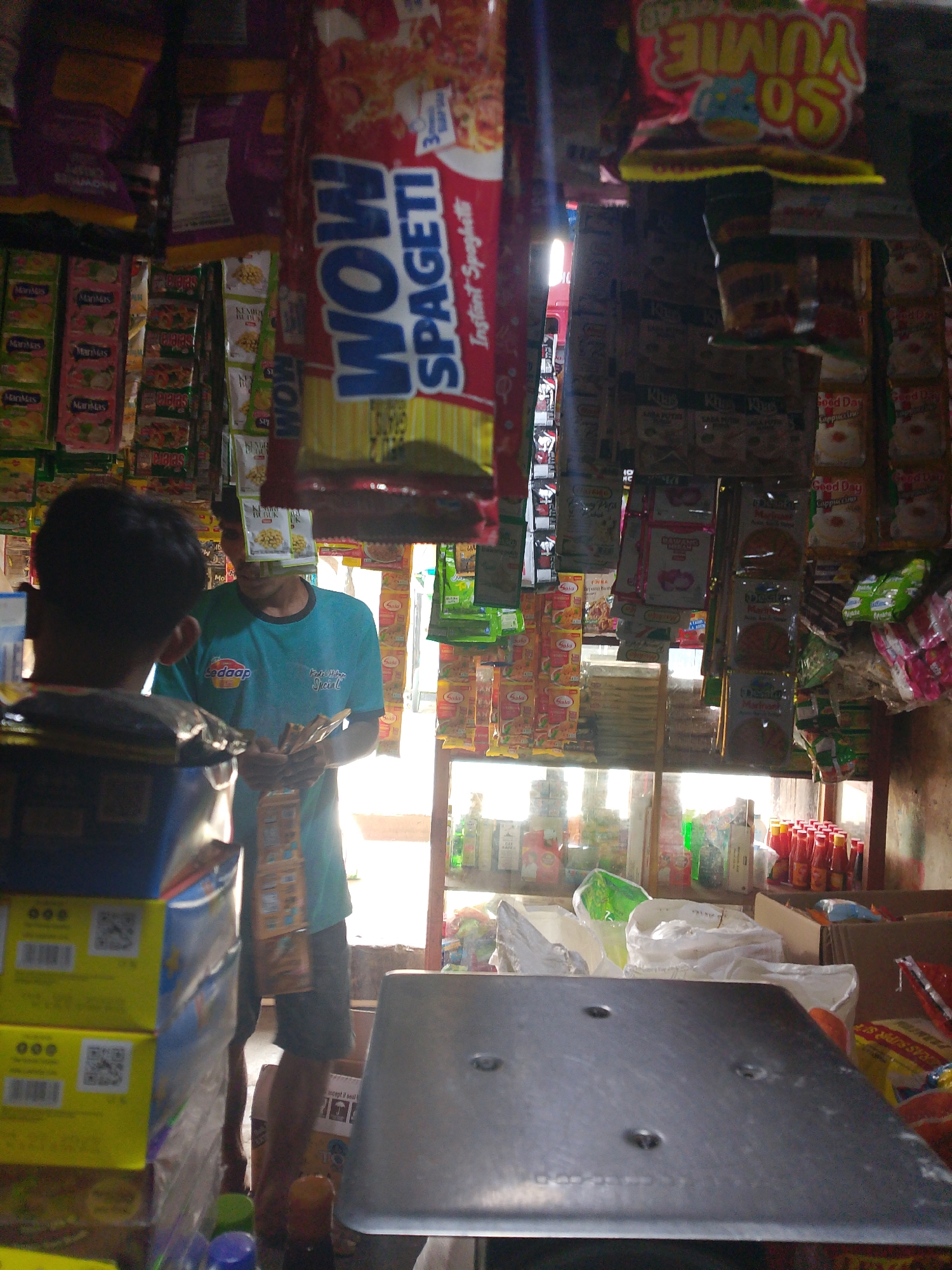The image depicts the interior of a small, cluttered convenience store in Palembang, Indonesia, likely in a residential area given the local products and the informal setup. In the foreground, the back of a person is visible, facing towards the store's interior. In the middle ground, a store attendant wearing a teal t-shirt is interacting with a customer whose back is to the camera. The attendant is holding several small packages, possibly food items or snacks, and appears to be in the process of a transaction. The store is densely packed with various packaged goods, including instant noodles, snacks, and other pantry staples, displayed on shelves and hanging from the ceiling. Products from local Indonesian brands are prominently featured. The lighting suggests a mix of natural light coming from the back of the store and possibly artificial lighting within. The overall impression is one of a typical neighborhood "warung" or small retail outlet serving the local community.