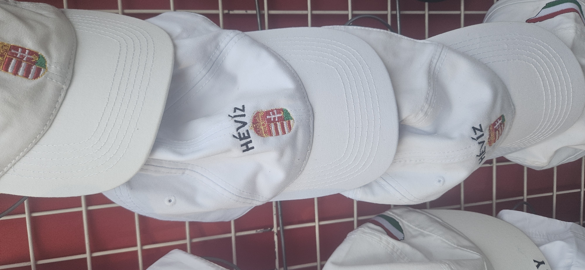 A stack of white baseball caps is displayed on a red grid rack, likely in a souvenir shop in Hévíz, Hungary. The caps are white and embroidered with various motifs. The cap at the top of the stack has a crest with red and white stripes and a green crown. The cap below it features the word "HÉVÍZ" in black text above a similar crest. Further down, another cap shows a portion of the Hungarian flag colors (red, white, and green) on its side. The caps are arranged in a vertical fashion, with the brim of one cap resting on the crown of the one below it. The background consists of a red metal grid, characteristic of market stalls or retail displays. There are no people visible in the image.
