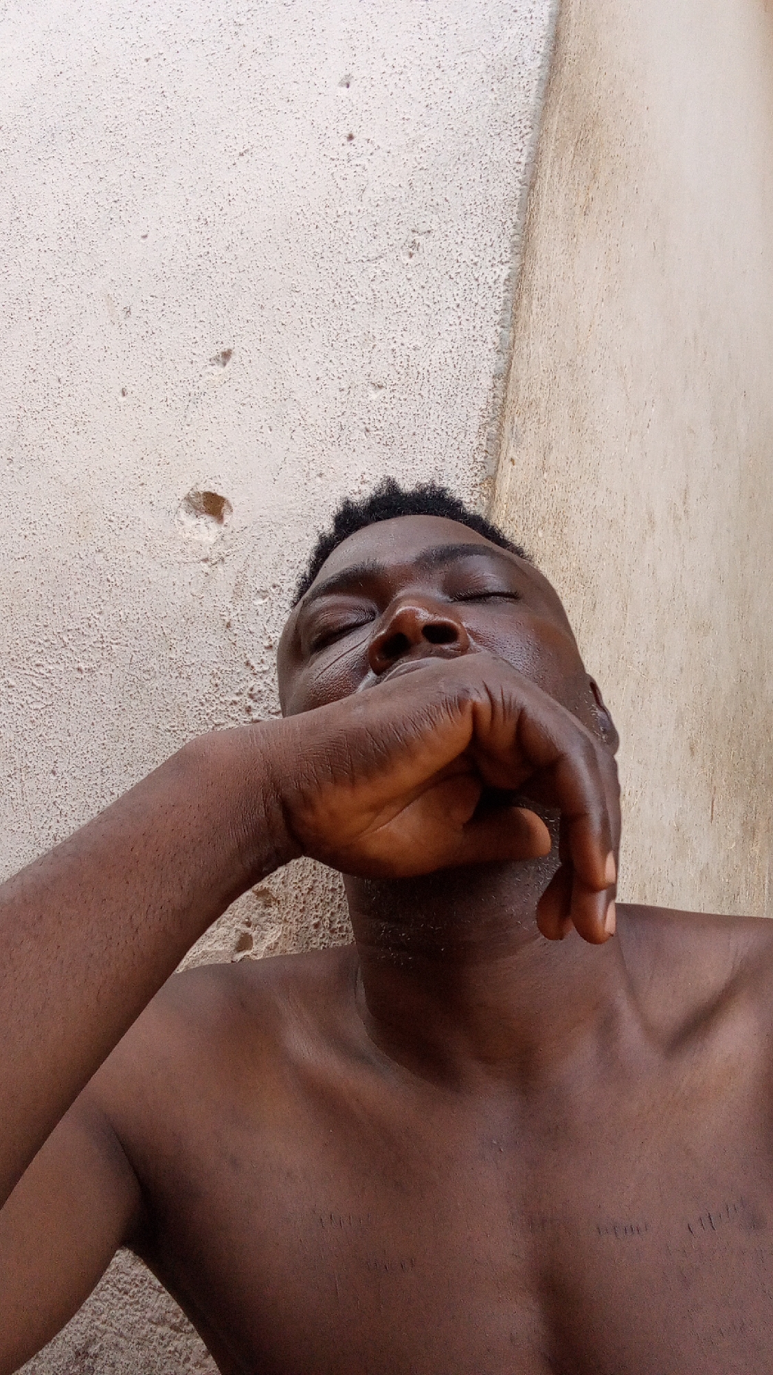 A close-up image depicts a dark-skinned man with a shaved head lying down, his eyes closed and his right hand covering his mouth. The background shows a textured wall that appears to be outdoors. The location is Unknown, Unknown.