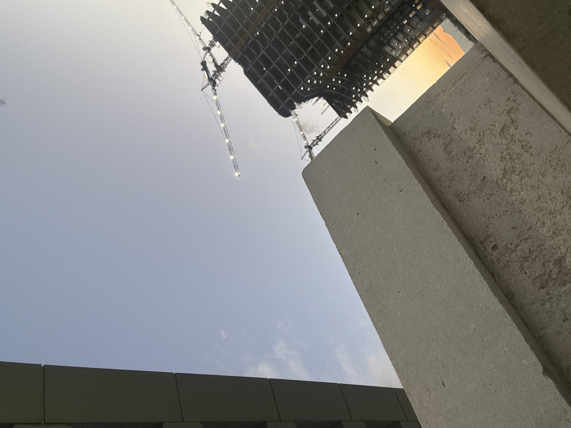 The image presents a low-angle, upward-looking view of a large-scale construction site in Yerevan, Armenia. The scene is dominated by a clear sky, transitioning from a light blue with faint clouds in the lower left to a brighter, yellowish-orange glow in the upper right, suggesting either sunrise or sunset.
In the upper portion of the frame, the dark, intricate underside of a massive structure, possibly a building or stadium roof under construction, is prominent. It features a grid-like pattern with numerous small, bright lights embedded throughout. Several large construction cranes with lights illuminating their long booms are visible, extending upwards from the left and right sides, indicating active building work.
In the foreground, on the right, rough-textured, light gray concrete pillars or sections of a building facade diagonally cut into the frame. A dark, horizontal structure forms the bottom edge on the left side. The overall composition highlights the industrial scale of the development and the intricate structural elements from an uncommon perspective. There are no people visible, and no discernible text.