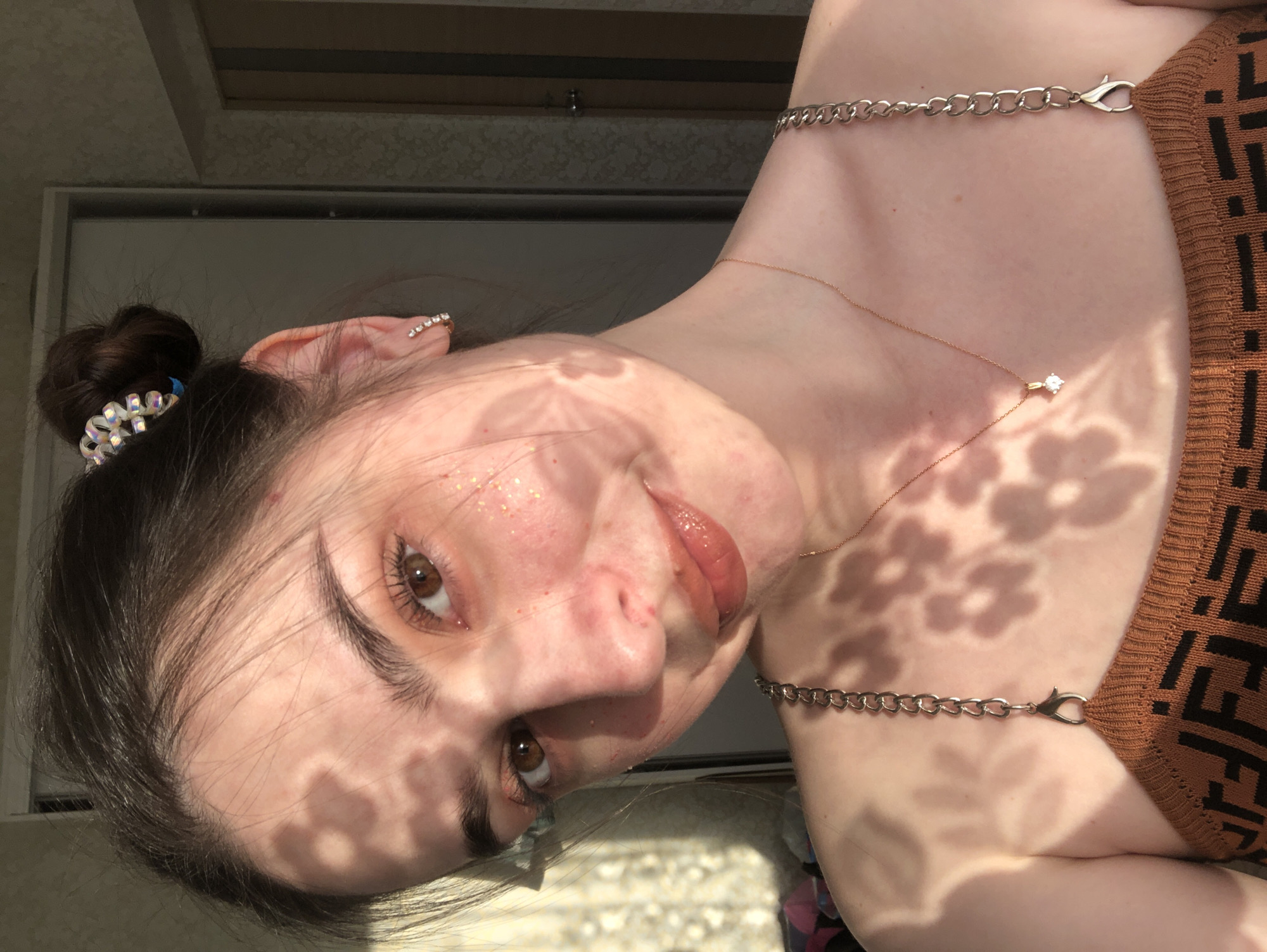 A young woman is posing for a photo. She has brown eyes and dark brown hair pulled back into a bun adorned with a sparkling hair accessory. She is wearing a brown top with a geometric pattern and a delicate gold necklace with a small diamond pendant. A silver chain-link strap of the garment is visible on her shoulder. The face is partially covered by shadows in the shape of flowers. She has fair skin and light freckles or glitter around her eyes. The background appears to be an indoor setting with a door or window frame visible behind her. Her lips are slightly parted as if posing for the camera, and her head is tilted towards the lens. There is no information about the location being in Noviy, Russia.