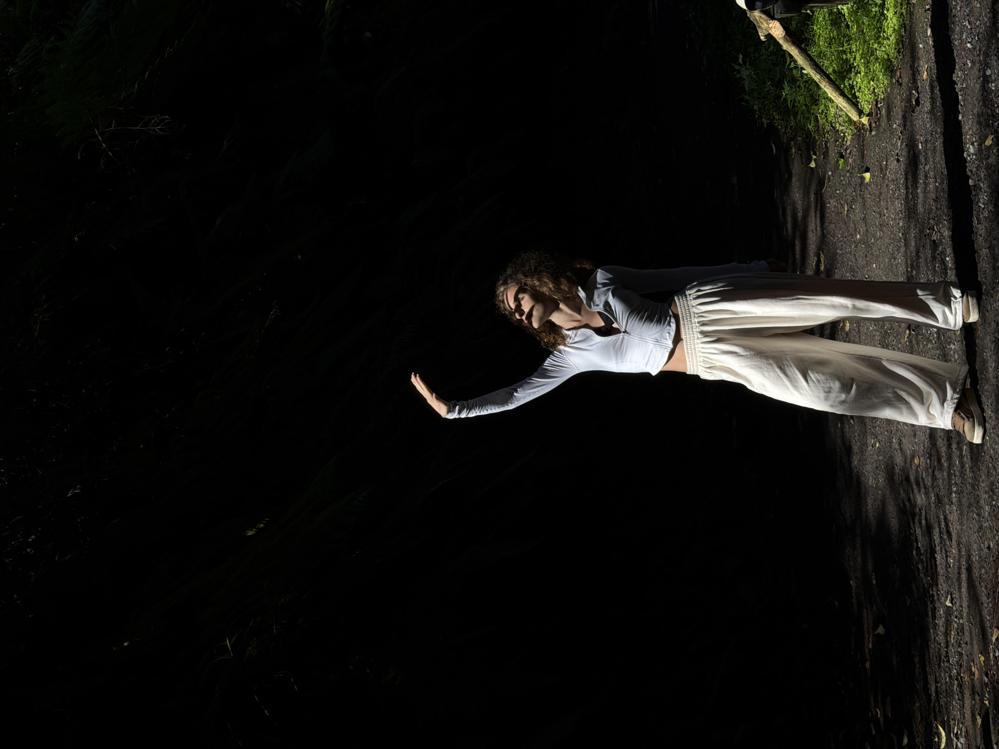 A young woman with curly brown hair stands in a dimly lit forest, her body angled to the left and her right arm raised towards the sky. She is wearing a white zip-up long-sleeved shirt and cream-colored wide-leg pants. Sunlight falls on her face and torso, casting shadows that obscure her eyes. The ground beneath her is a dirt path covered with small stones and fallen leaves. To the right, a thick patch of green grass is visible, with a fallen tree branch intersecting the path and grass. The background is a deep shadow, with faint suggestions of foliage. The image appears to have been taken in the vicinity of Penebel, Indonesia, given the lush tropical vegetation.