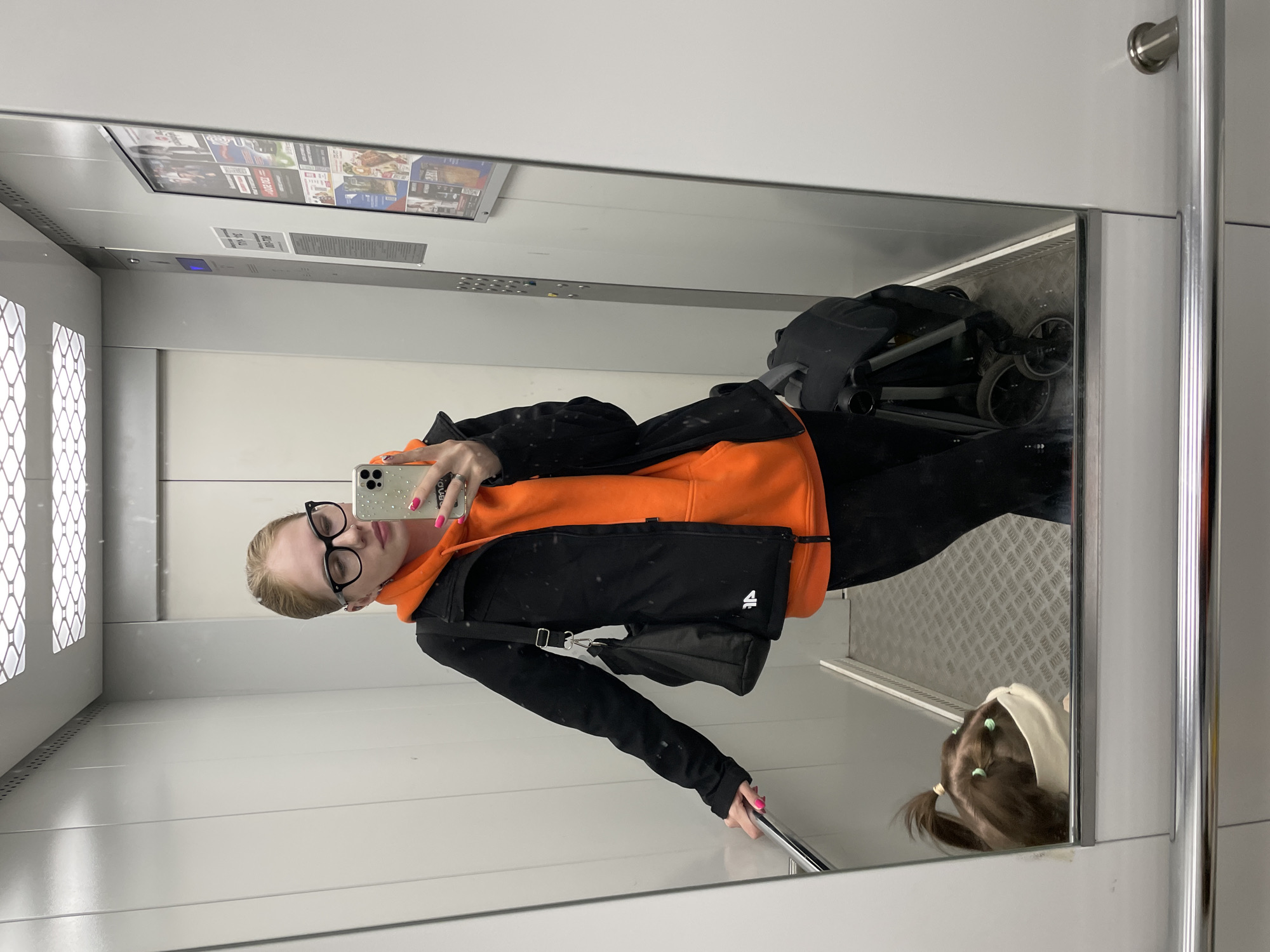 A young woman takes a mirror selfie in an elevator. She stands vertically, looking directly at her smartphone, which she holds up to capture her reflection. She has light-colored hair pulled back, wears black-rimmed glasses, an orange hoodie, and a black jacket. Her nails are painted bright pink, and she wears a silver ring on her left hand. A black shoulder bag is visible on her left side. Her right hand rests on a chrome handrail.
The elevator interior features light-colored walls, possibly white or light grey, with a large mirror dominating one side. The floor, reflected in the mirror, appears to be made of textured metal, possibly diamond plate. Above the woman's head, an elevator control panel is visible, featuring buttons and a blue digital display. Further up, a ceiling-mounted display unit shows several posters or advertisements.
Reflected in the bottom right corner of the mirror is the back of a child's head, with brown hair styled in pigtails adorned with light green hair ties. Part of a light-colored hood or headwear is also visible on the child. Behind the woman and to her right, also reflected in the mirror, is a dark-colored baby stroller or pram, suggesting she is traveling with a child.
On the poster display unit, various images and some text are visible, including what looks like part of a red box with "2020" or "2021" and possibly the word "CLUB" or "KLIUB". Other posters show images of people, food, or products, typical of advertisements. The scene is brightly lit by artificial light. The overall setting is a functional, modern elevator in Izhevsk, Russia.